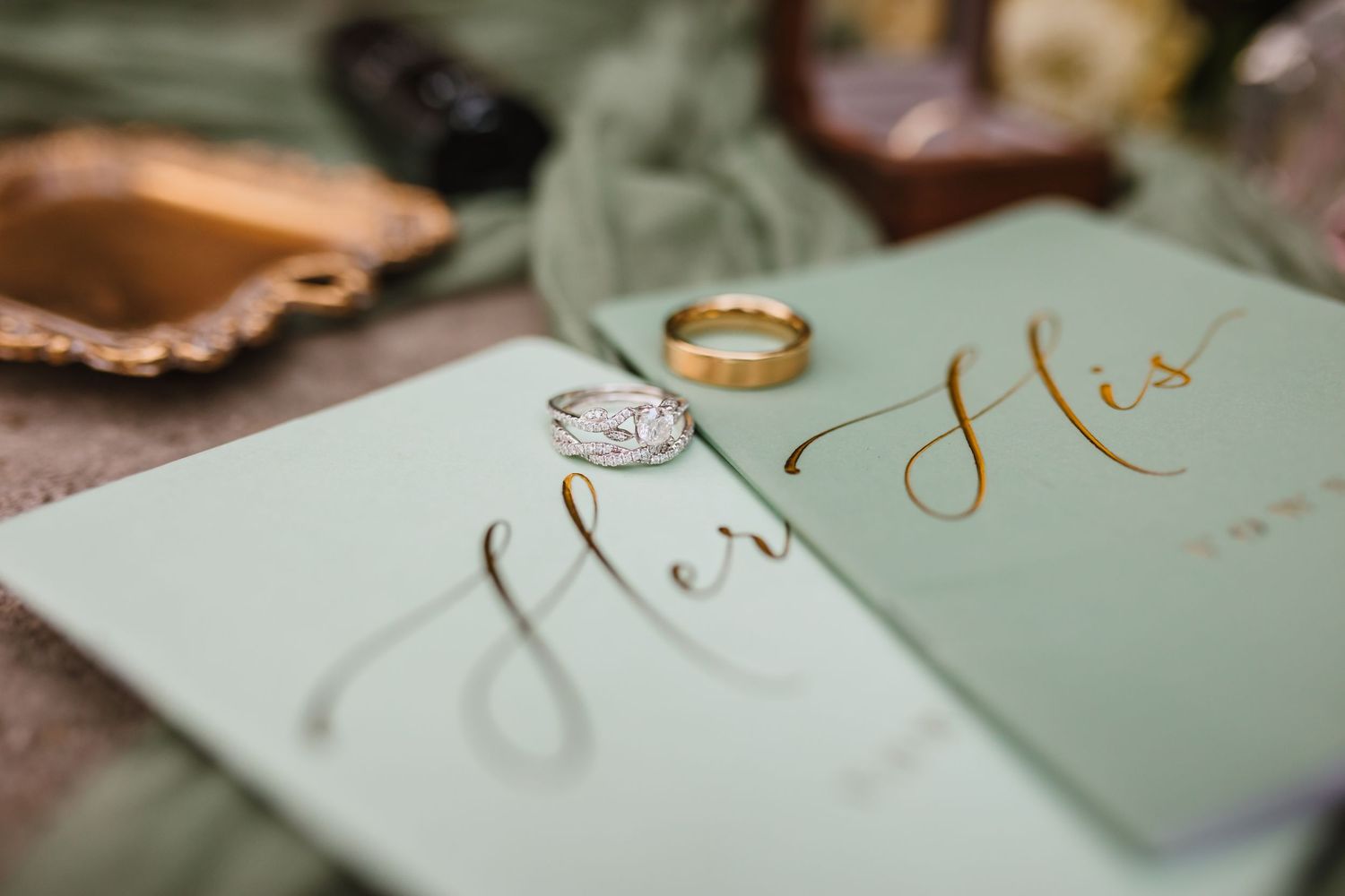 Wedding vow books labeled Her and His with gold rings on a sage green elegant stationery flat lay.