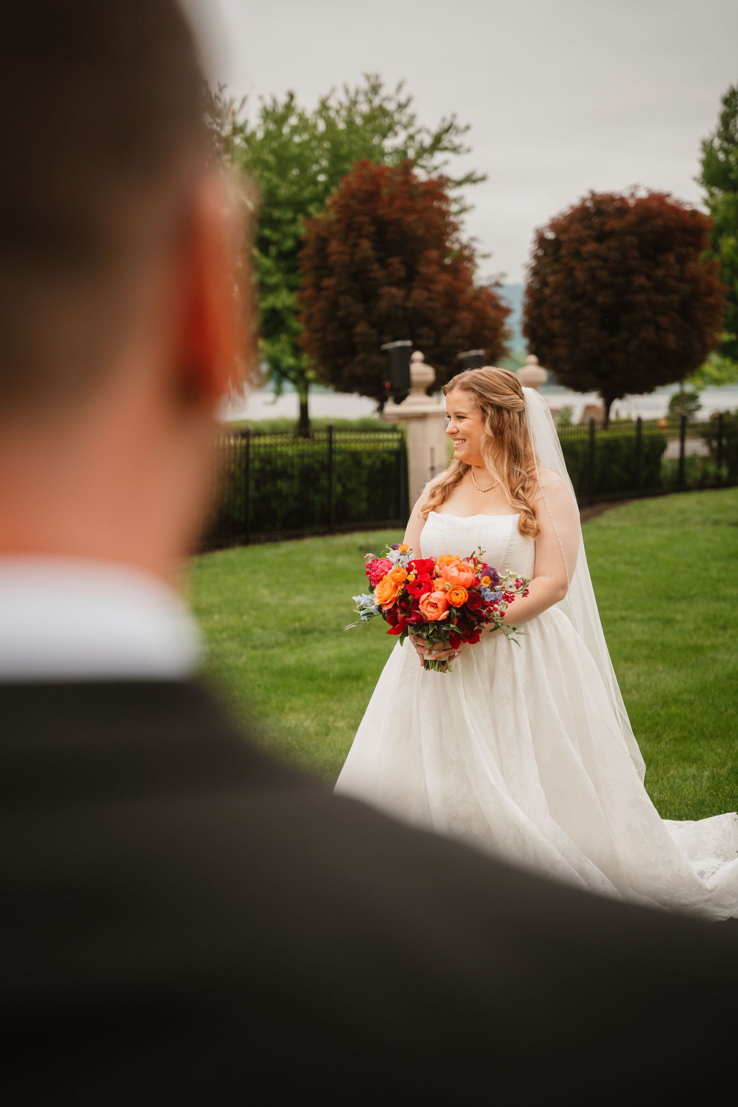 Smiling bride in white gown holds vibrant bouquet during first look with groom in garden.