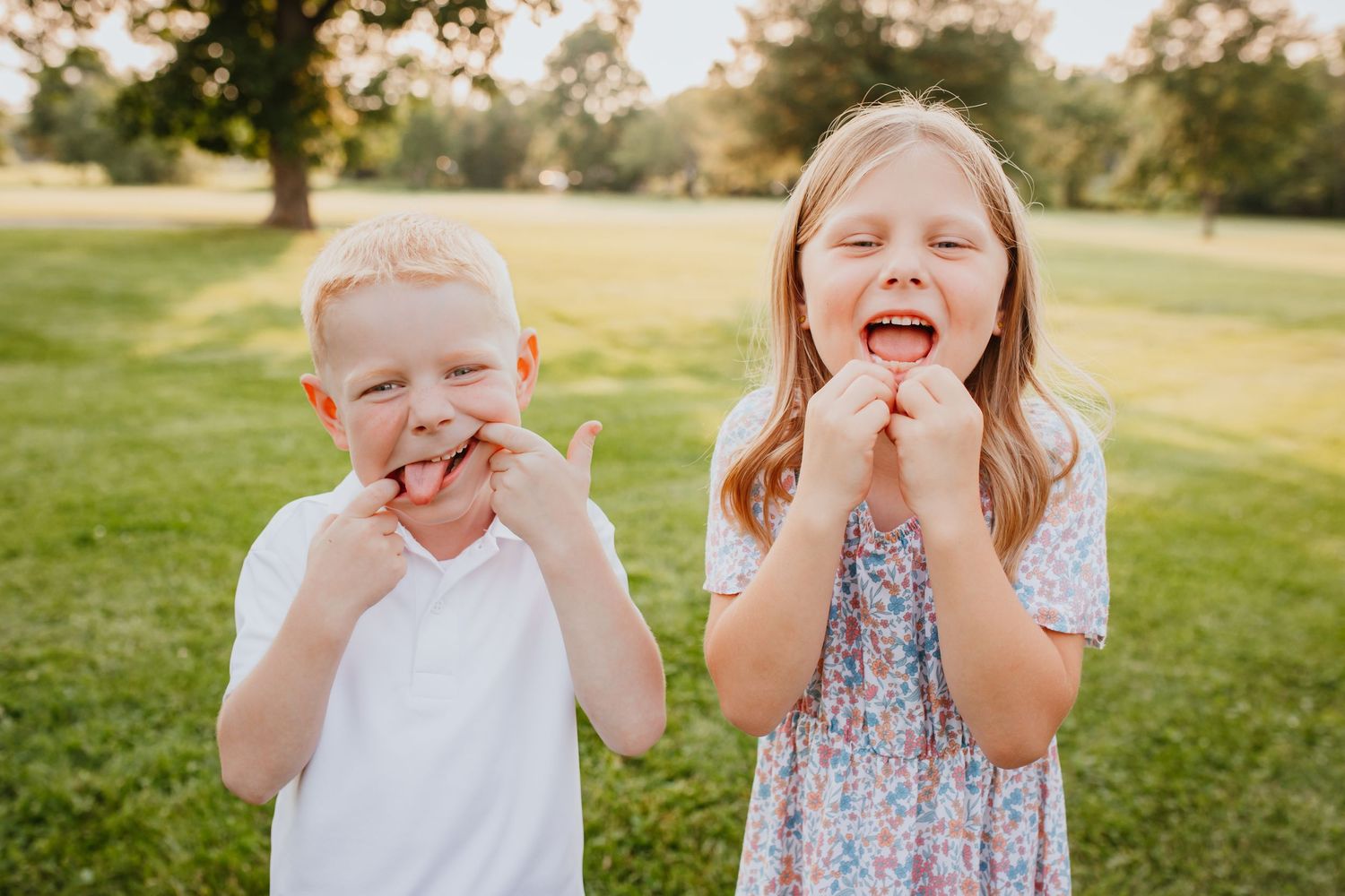 Two young siblings making silly faces and laughing together outdoors on a sunny summer day.