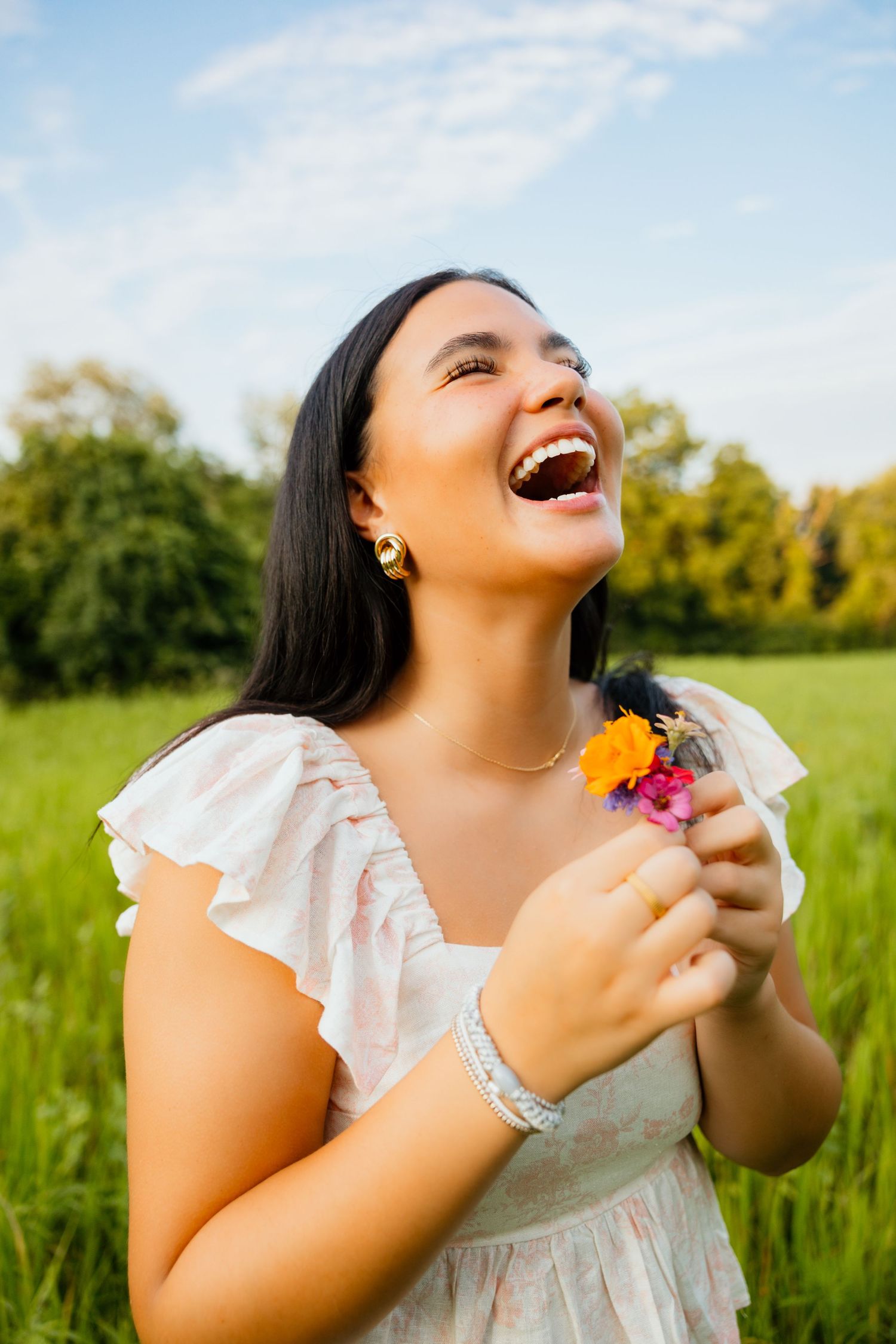 Joyful young woman with dark hair laughs while holding colorful wildflowers in a sunny green field.