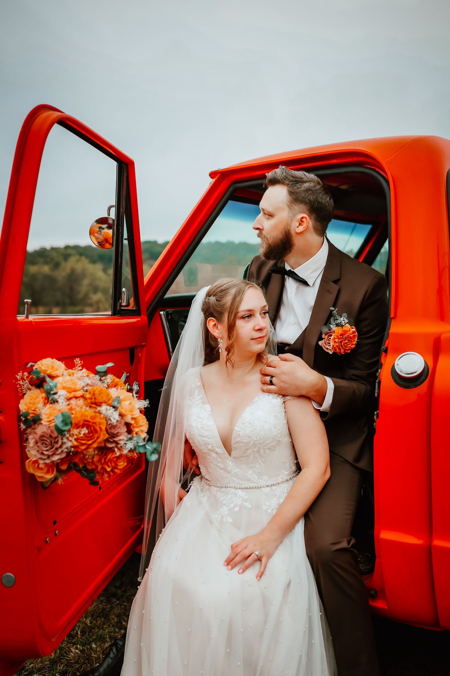 Bride and groom posing together by open door of classic red truck with fall floral bouquet.