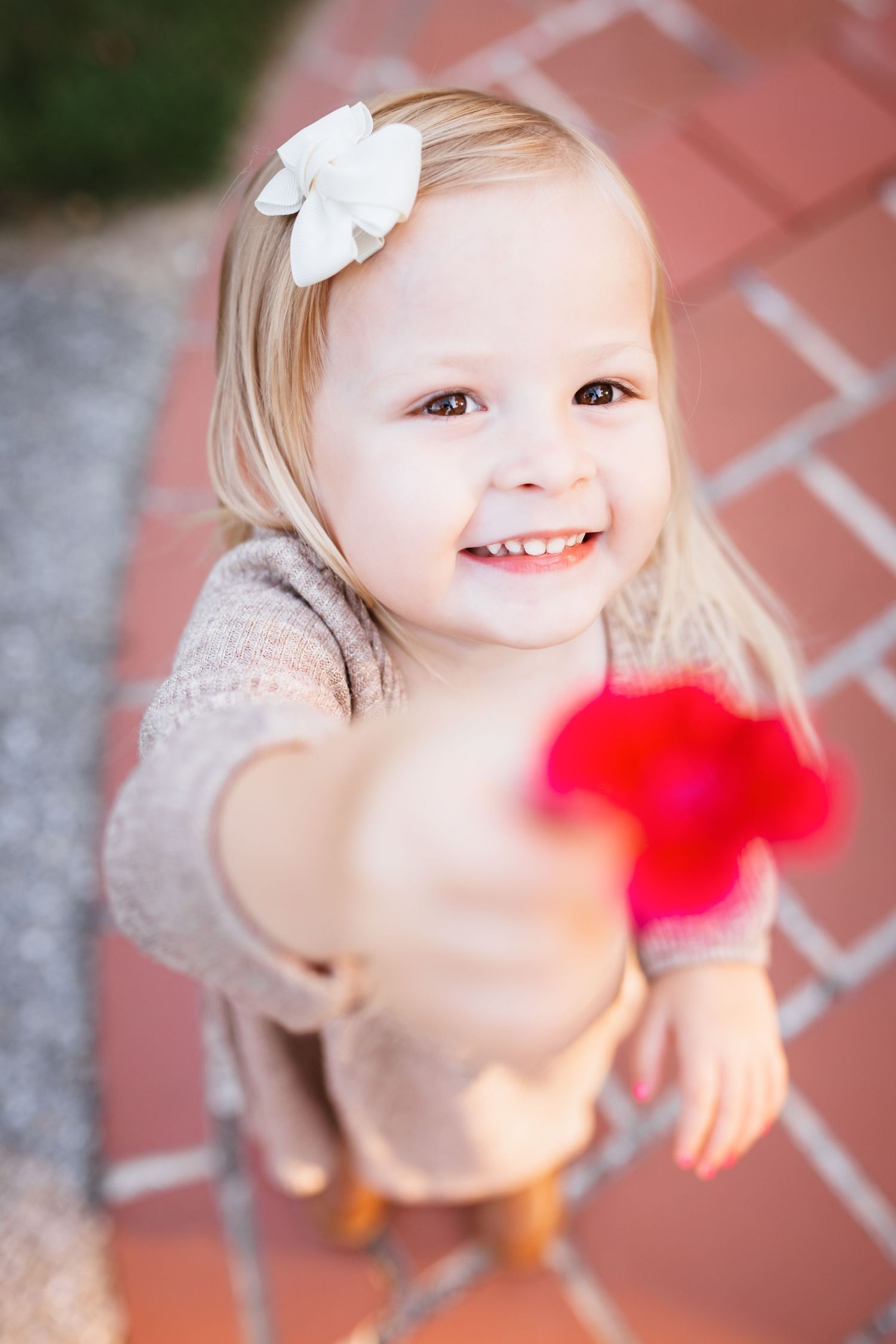 Cute smiling toddler girl with white bow holding red flower toward camera on brick path.