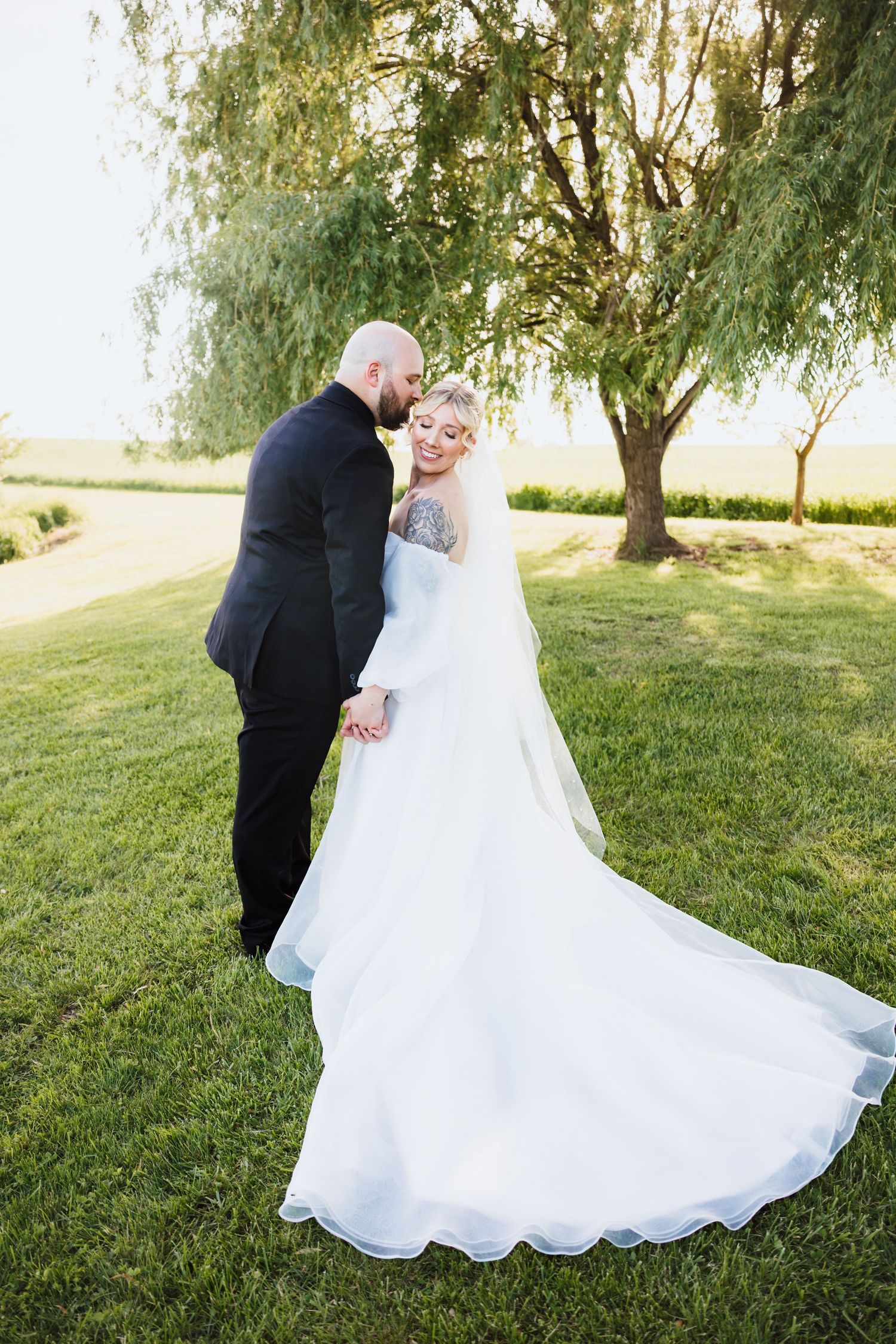 Bride in white gown with long train and groom in black suit share romantic moment under willow tree.