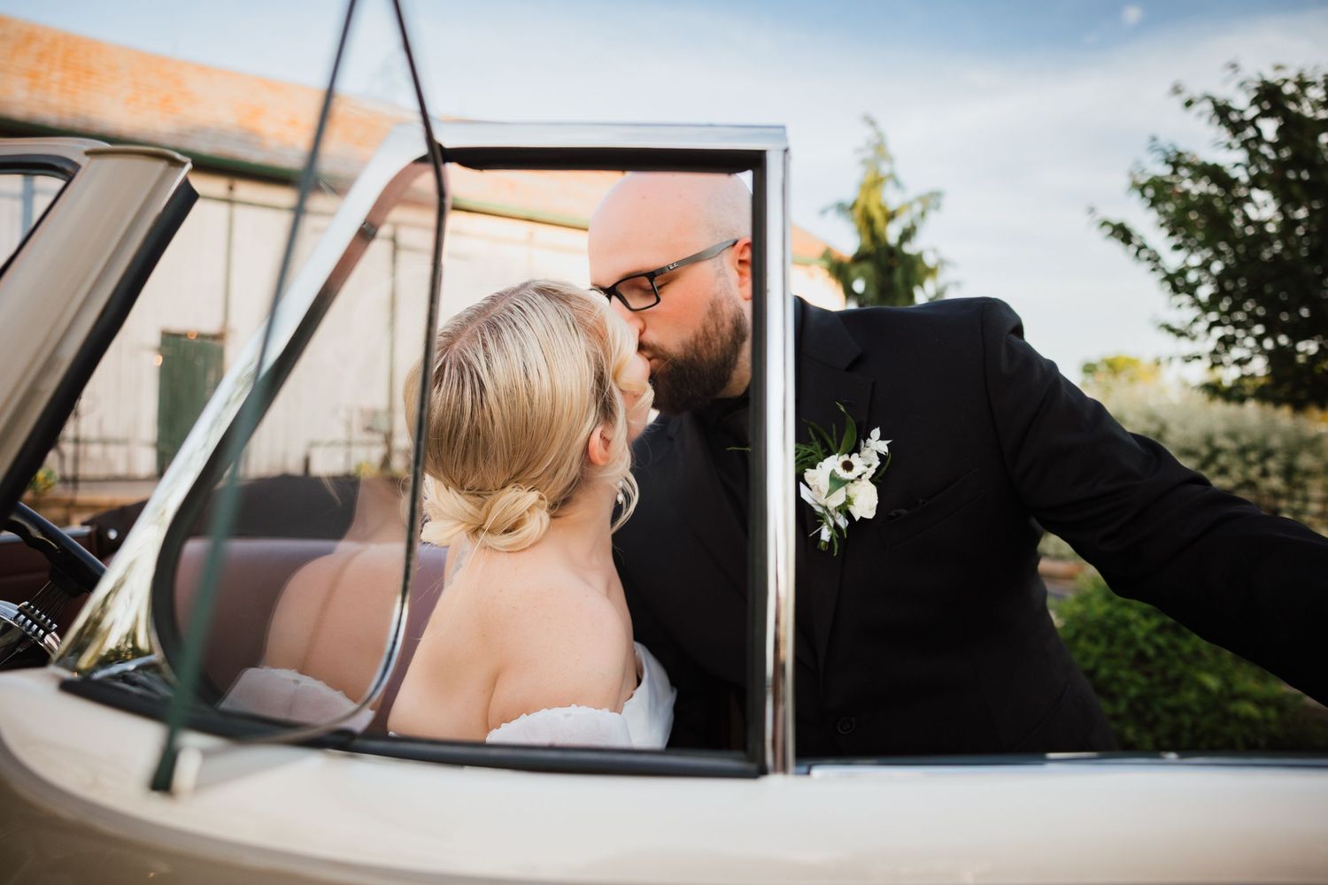 Groom kissing bride through vintage white convertible car window at outdoor wedding venue.