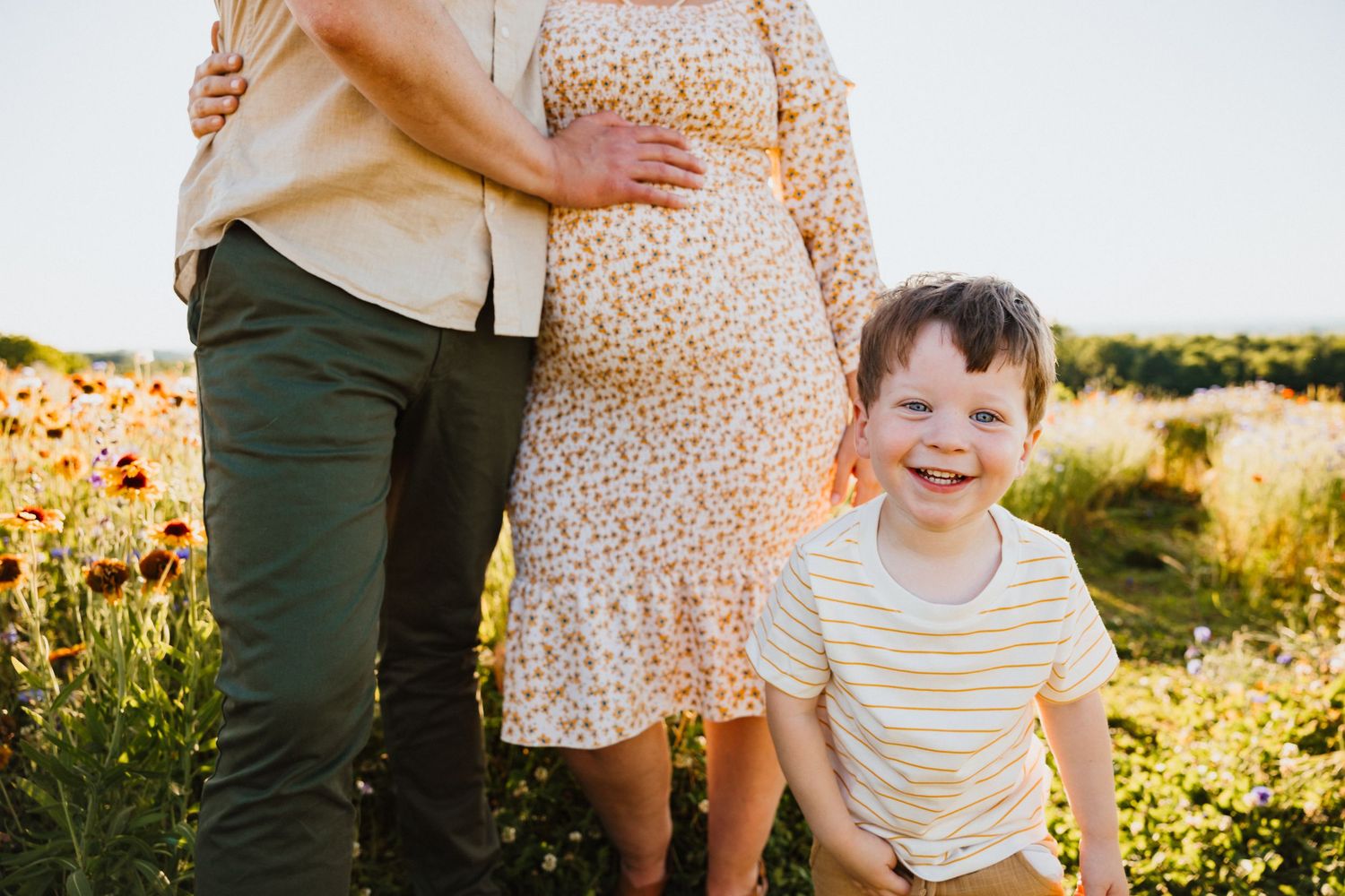 Smiling boy poses in front of pregnant mother and father in sunny wildflower field.