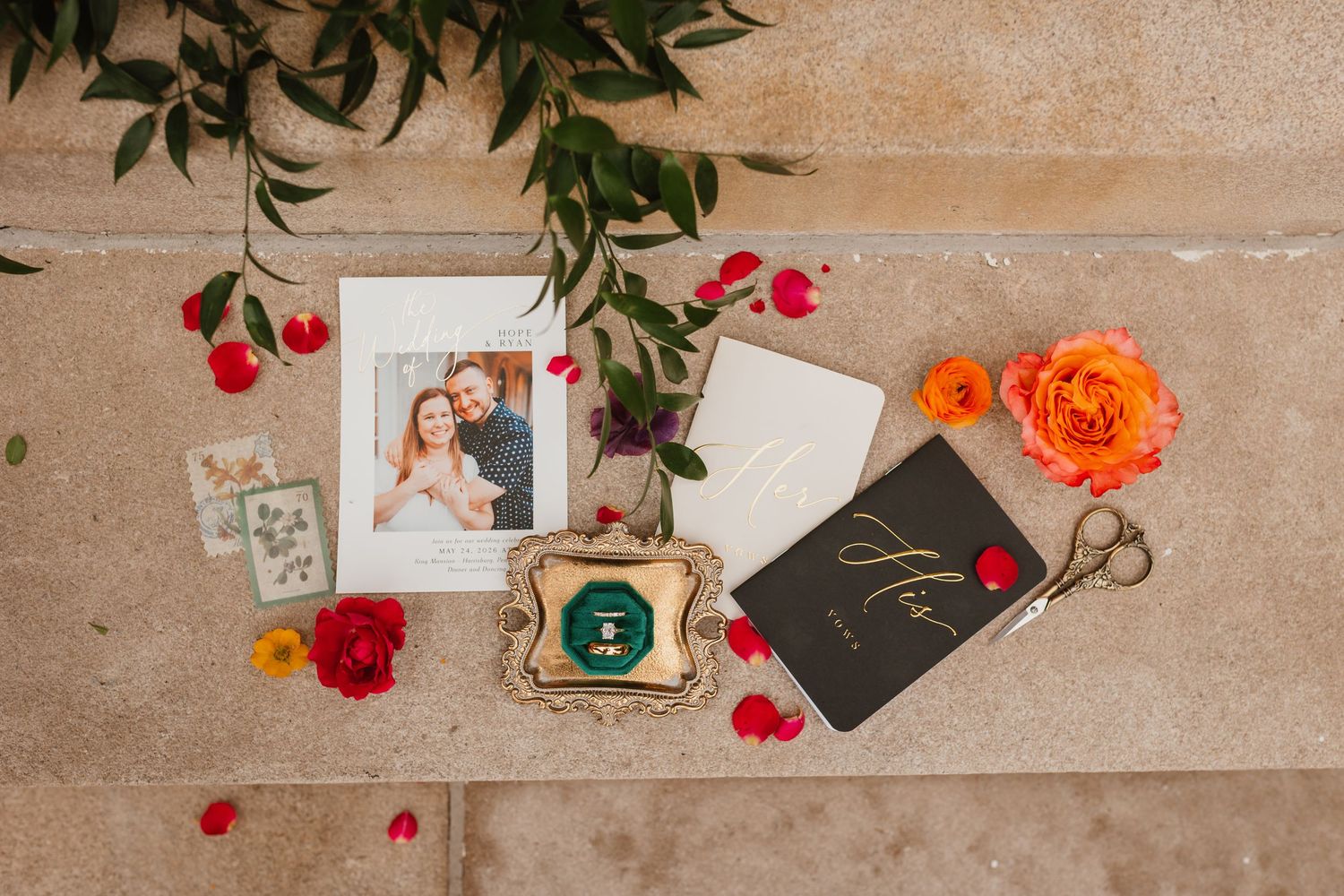 Wedding flat lay with couple photo, green velvet ring box, stationery, scissors, and colorful flowers.
