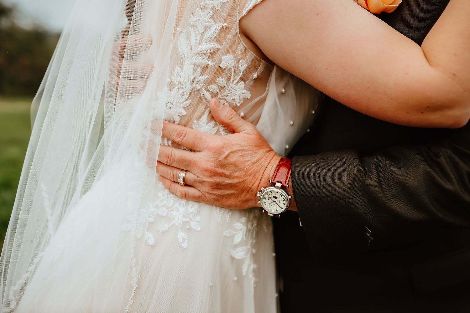 Close-up of bride and groom embracing showing lace wedding dress veil and groom watch detail.