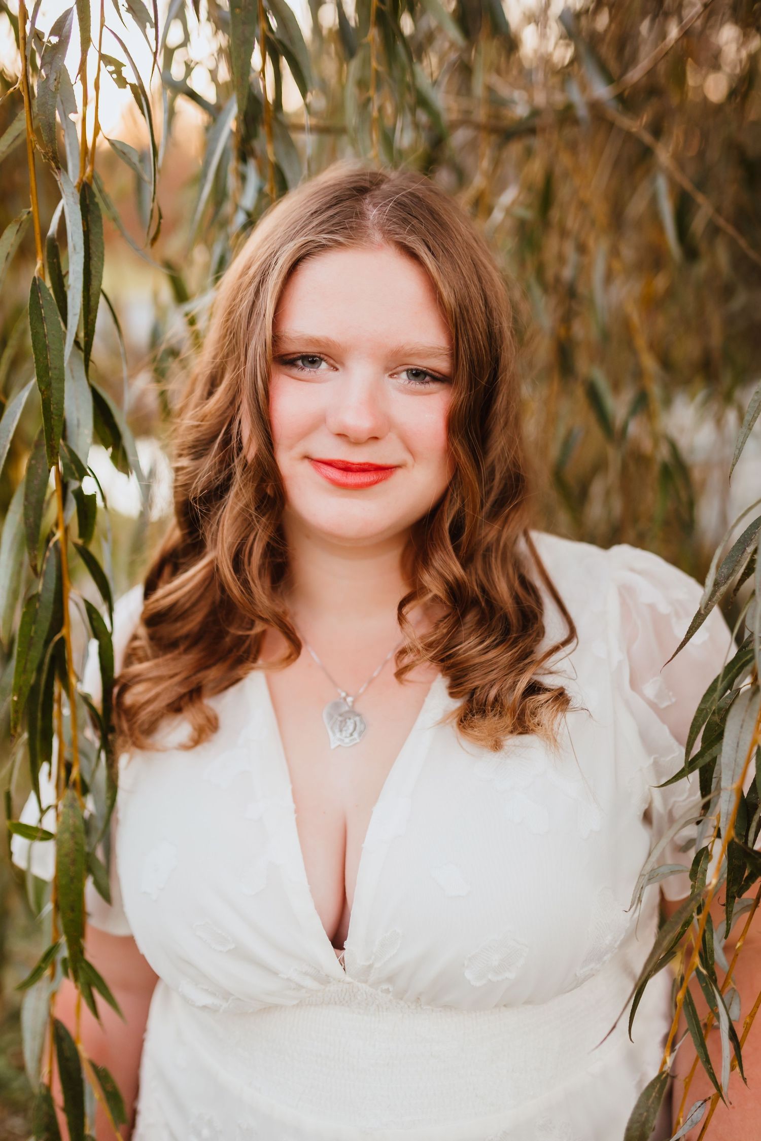 Young woman with auburn curls in white v-neck dress poses under weeping willow branches at sunset.