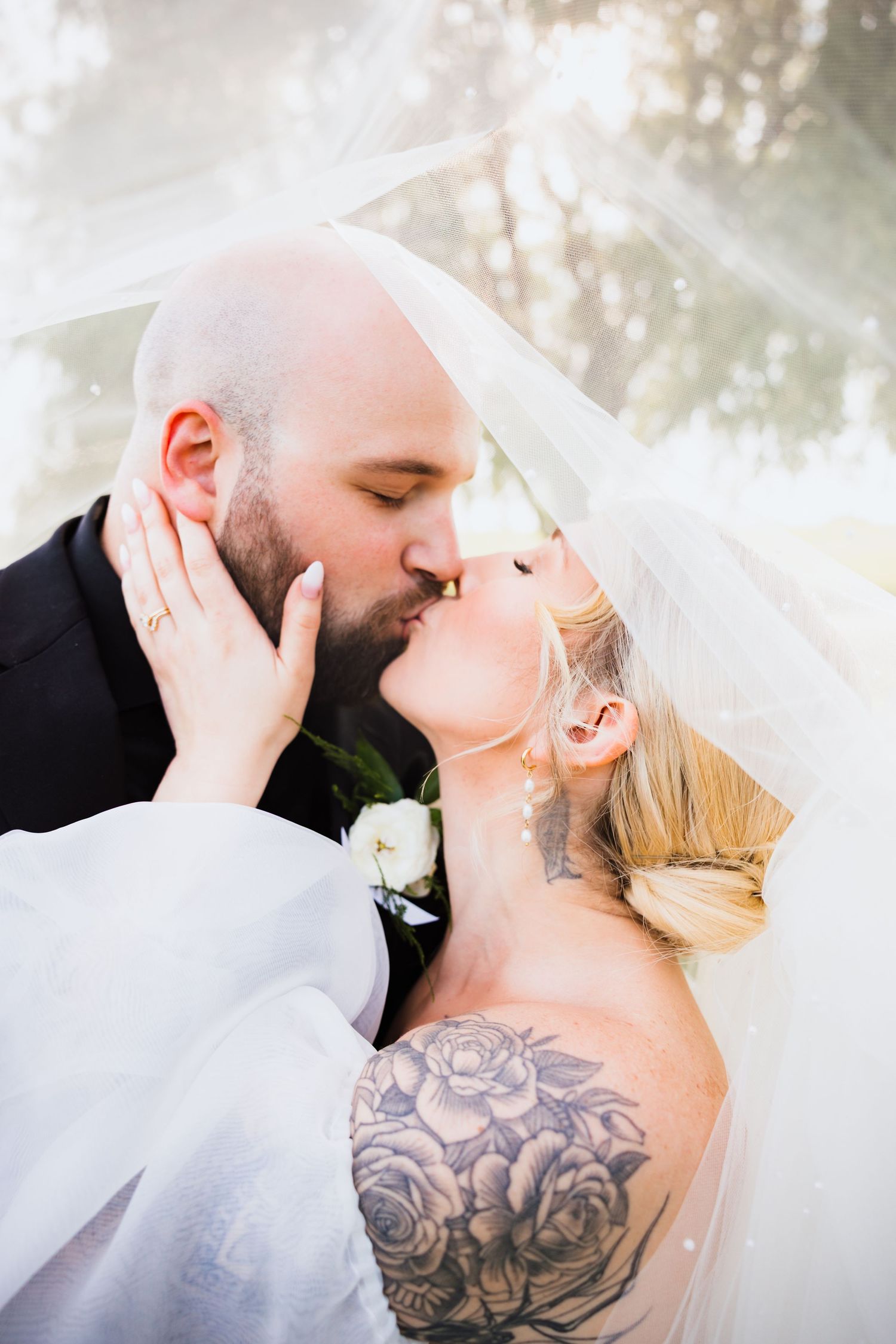 Close-up of bride and groom kissing under veil, bride showing rose tattoo on shoulder.