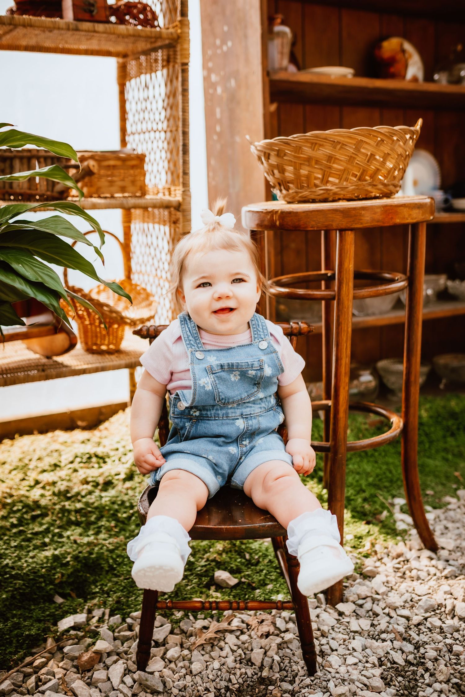 Happy baby girl in denim overalls sitting on a wooden chair surrounded by rustic wicker baskets outdoors.