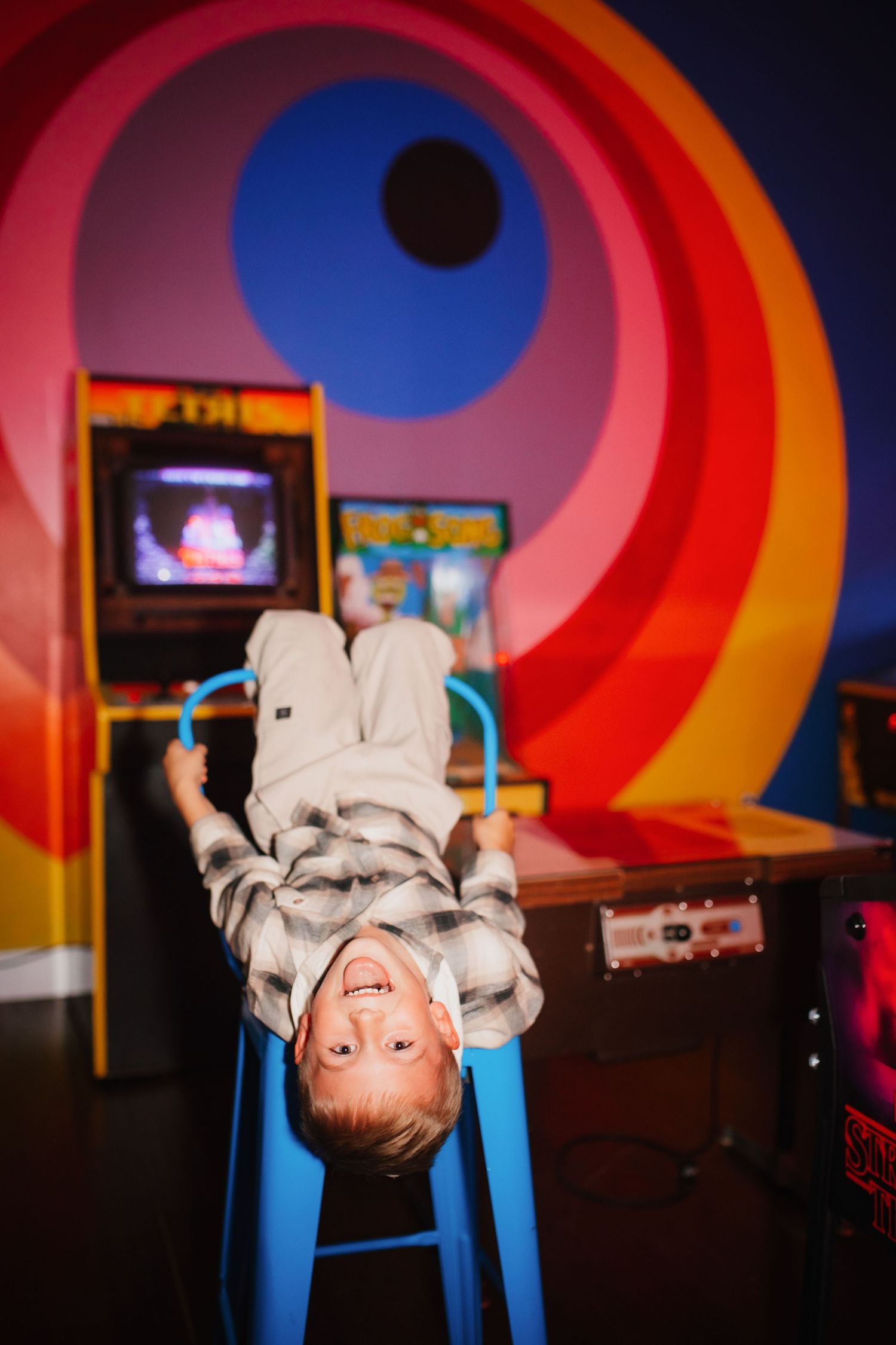 Happy boy hanging upside down on blue stool in colorful retro arcade game room.