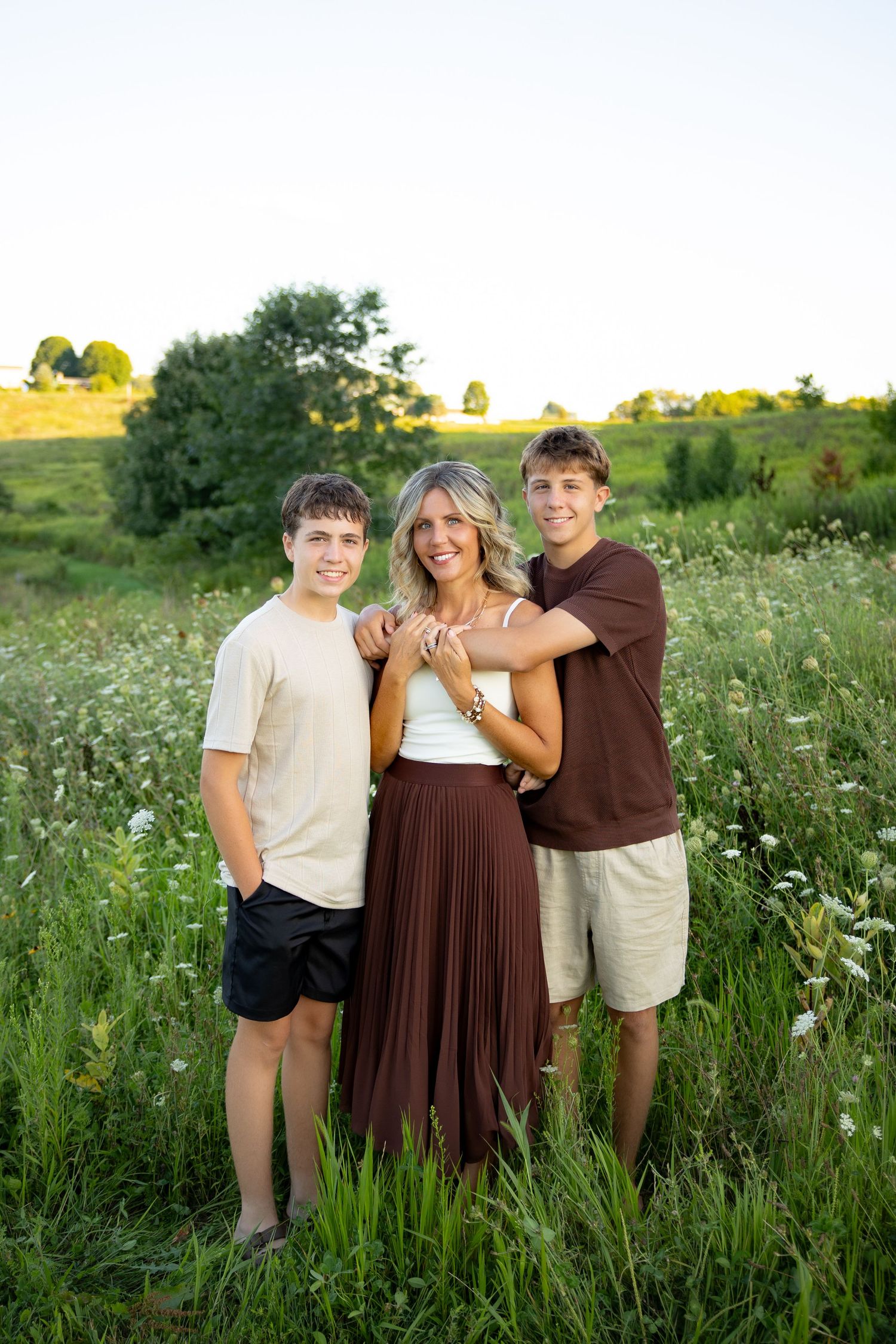 A smiling woman in a brown skirt poses with two teenage boys in a lush green field at golden hour.