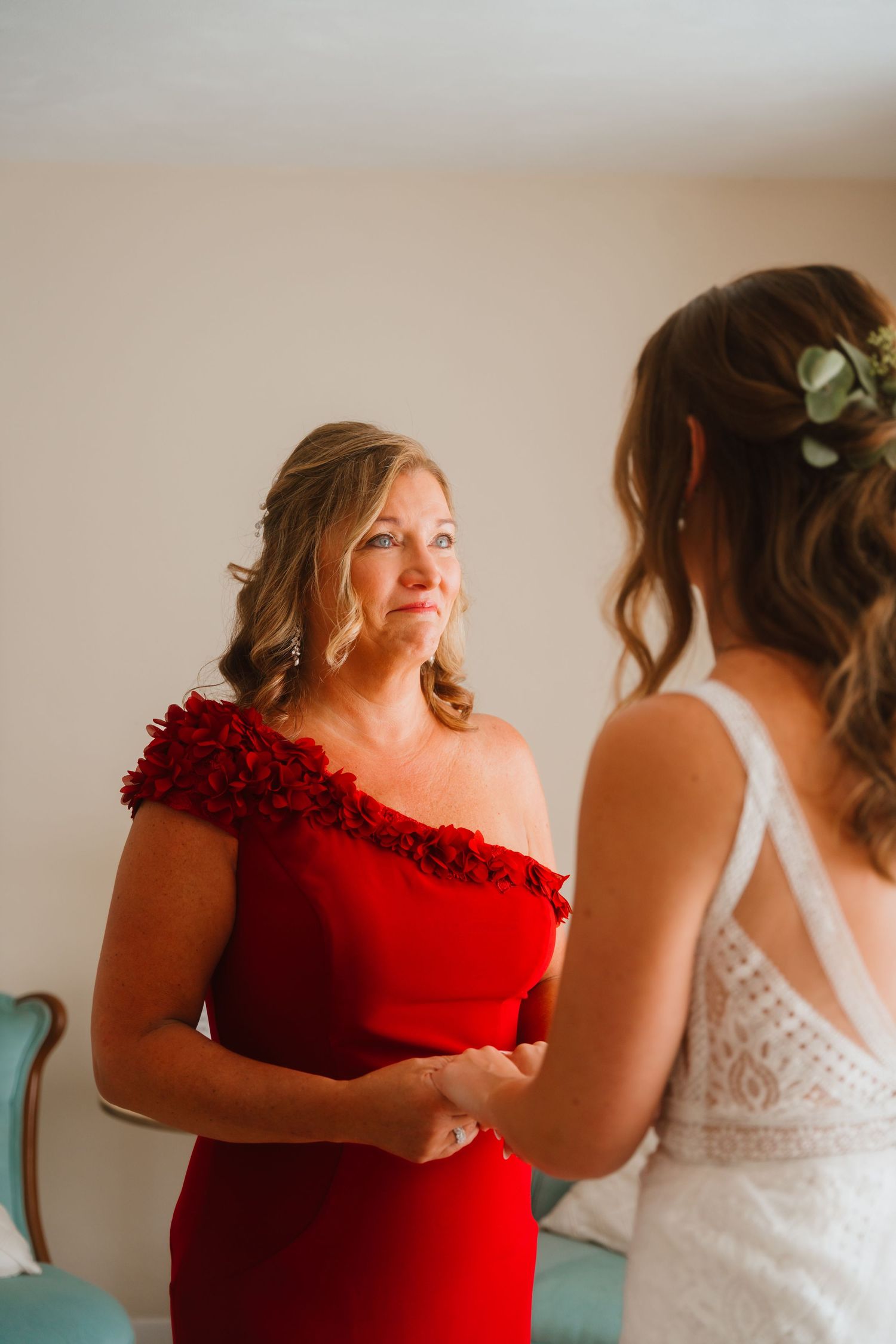 Mother in red dress holding hands with bride in white lace gown during wedding preparation.