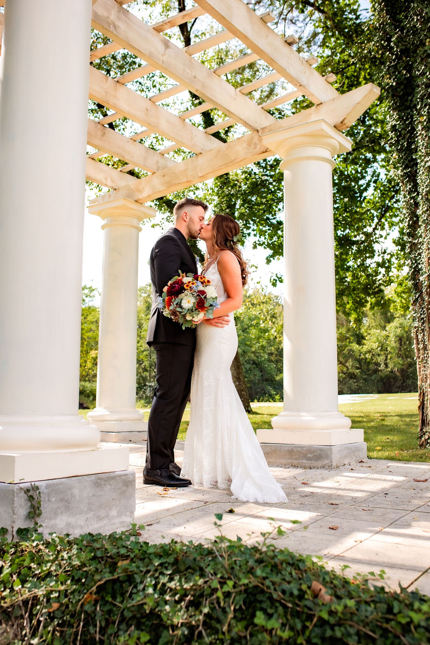 Bride and groom share a kiss under a white pergola surrounded by lush greenery at their outdoor wedding.