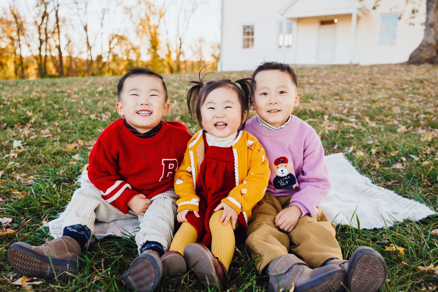 Three young Asian siblings sitting together on a blanket outdoors, smiling joyfully in autumn sunlight.