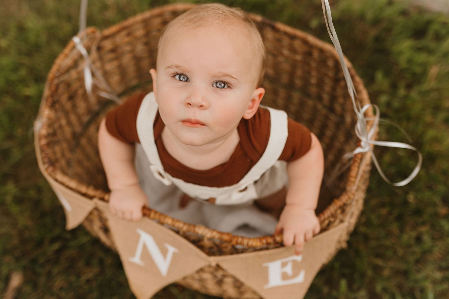 Blue-eyed baby in rust and white outfit sits in wicker basket with ONE burlap banner.