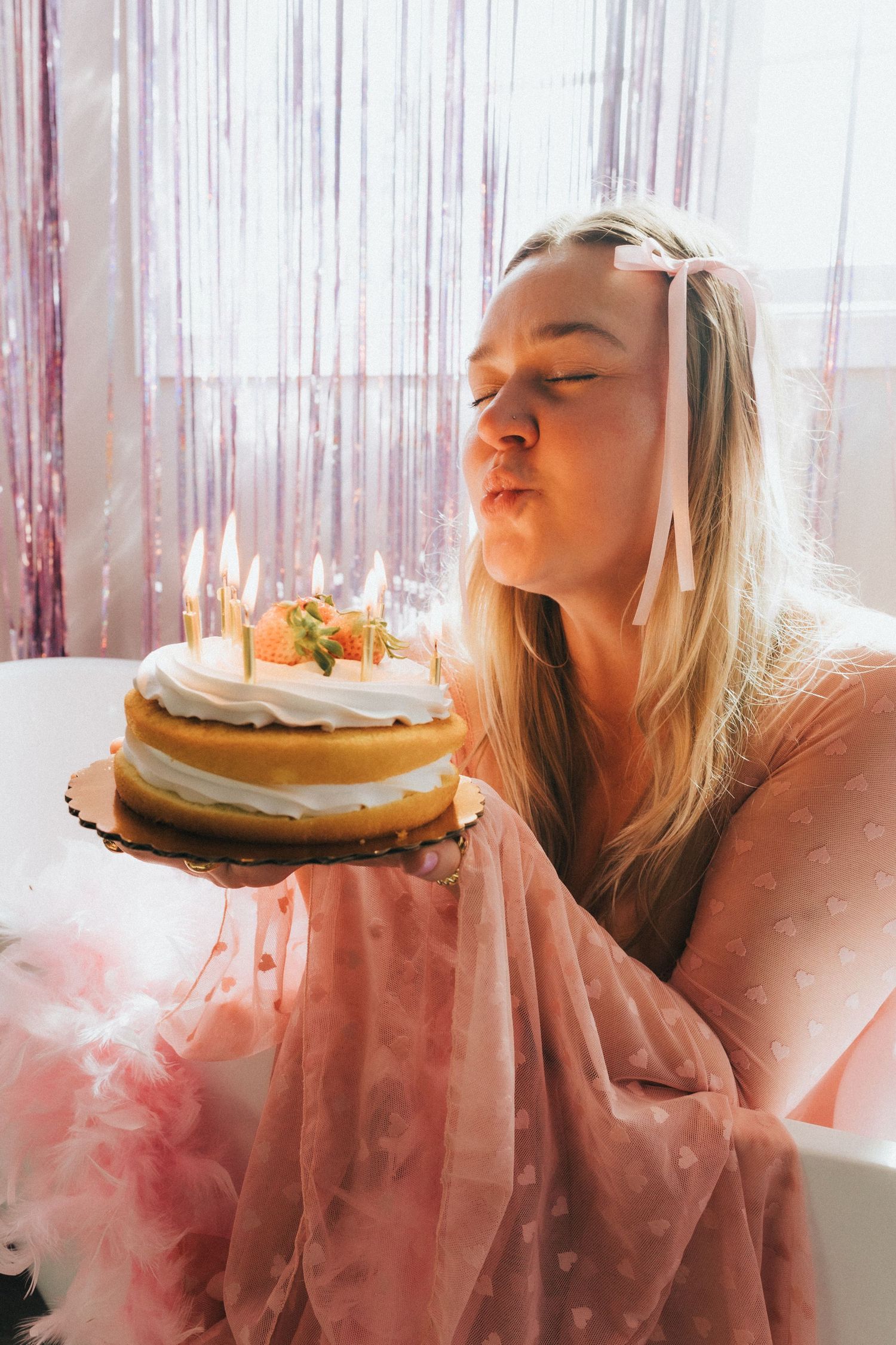Young blonde woman blows out candles on layered birthday cake with pink tinsel curtain background.