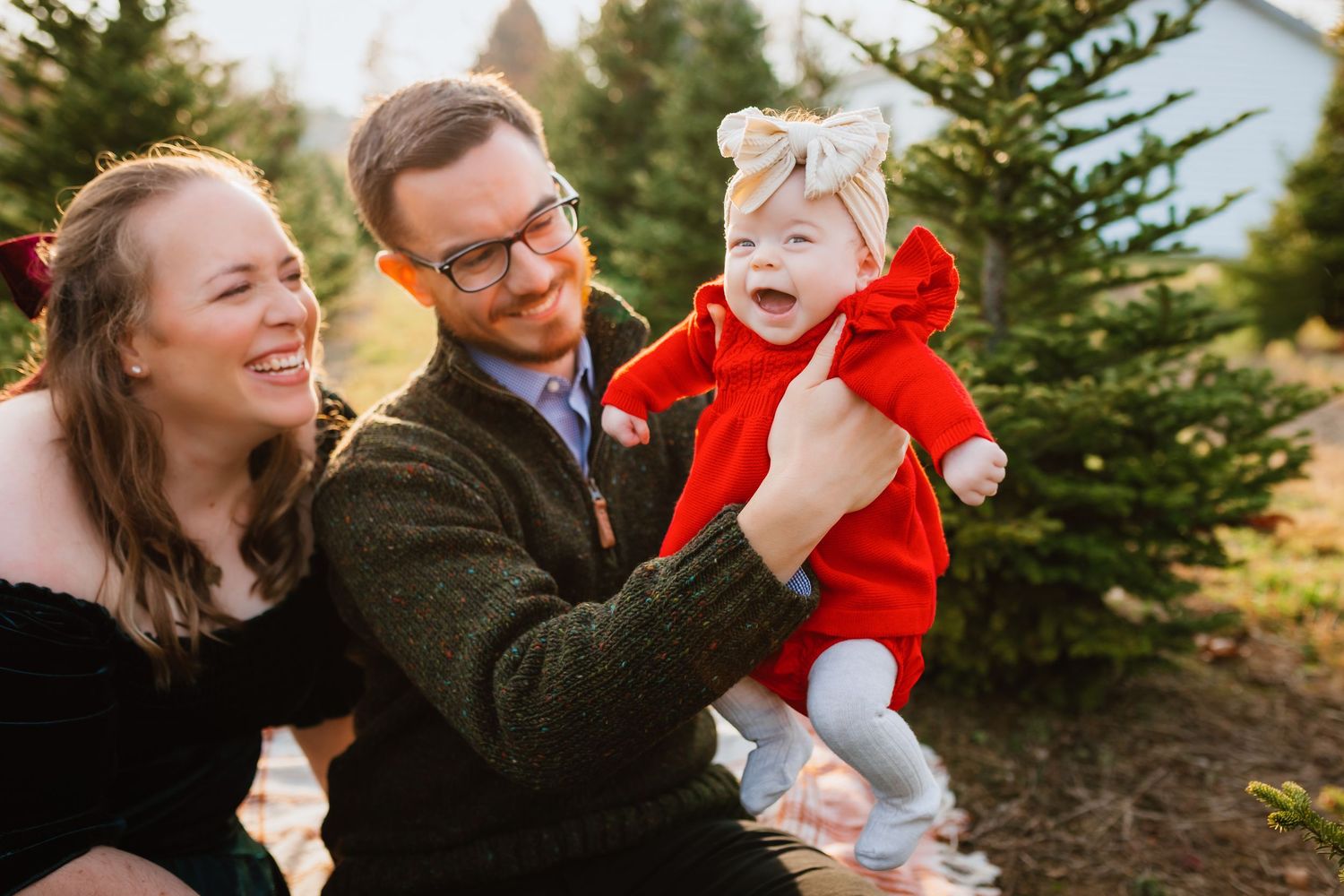 Happy parents laughing with baby girl in red outfit at Christmas tree farm during golden hour.