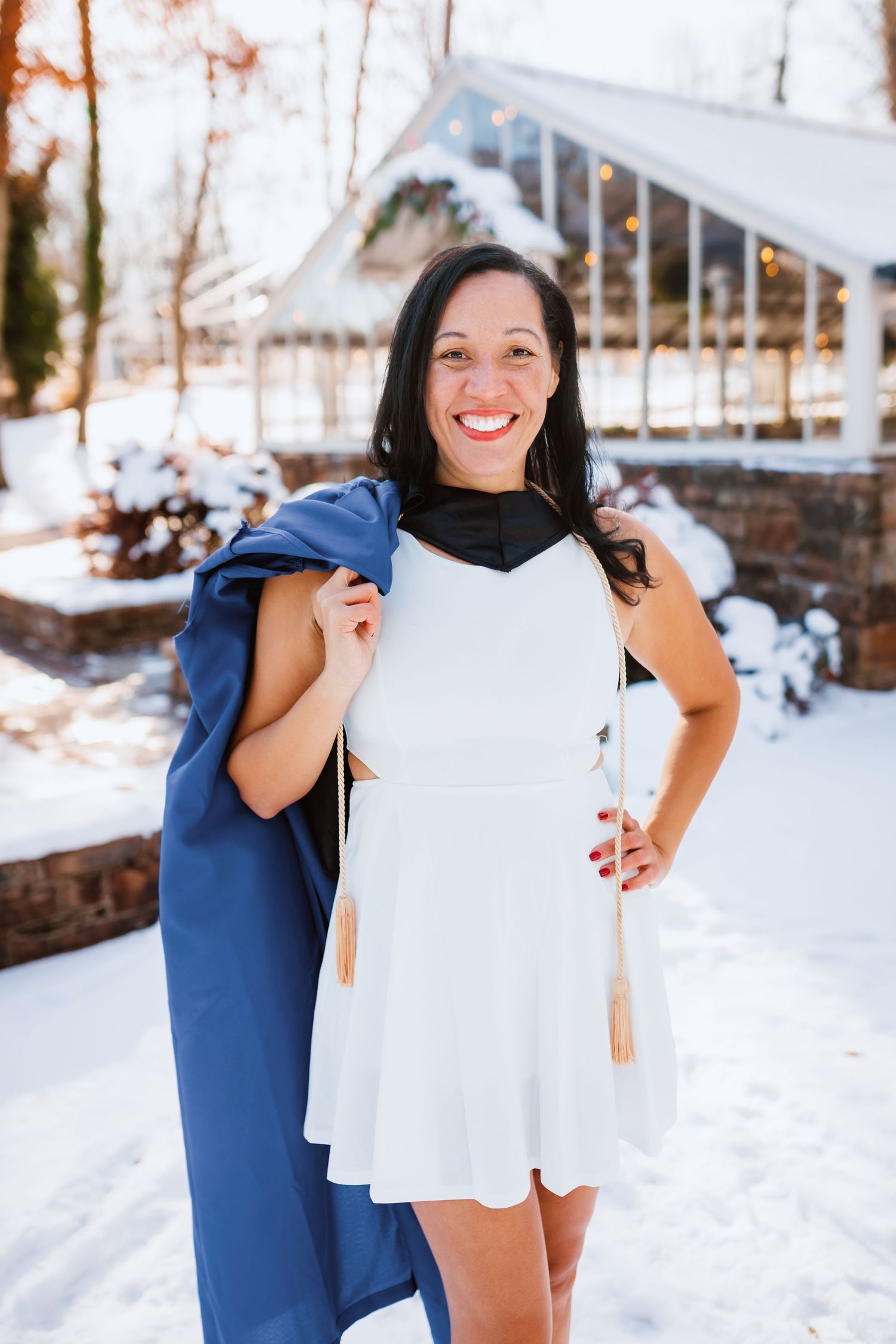 Smiling woman in white dress holds blue graduation gown over shoulder in winter setting.