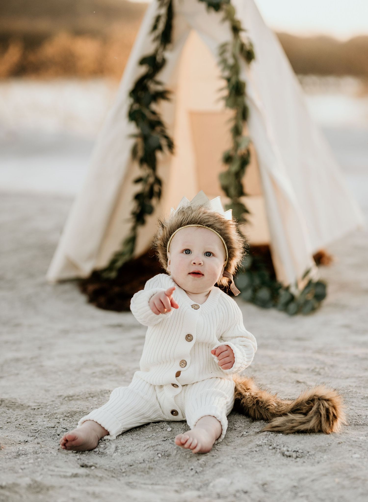 Baby in white ribbed outfit sitting on beach sand in front of boho teepee decorated with greenery.