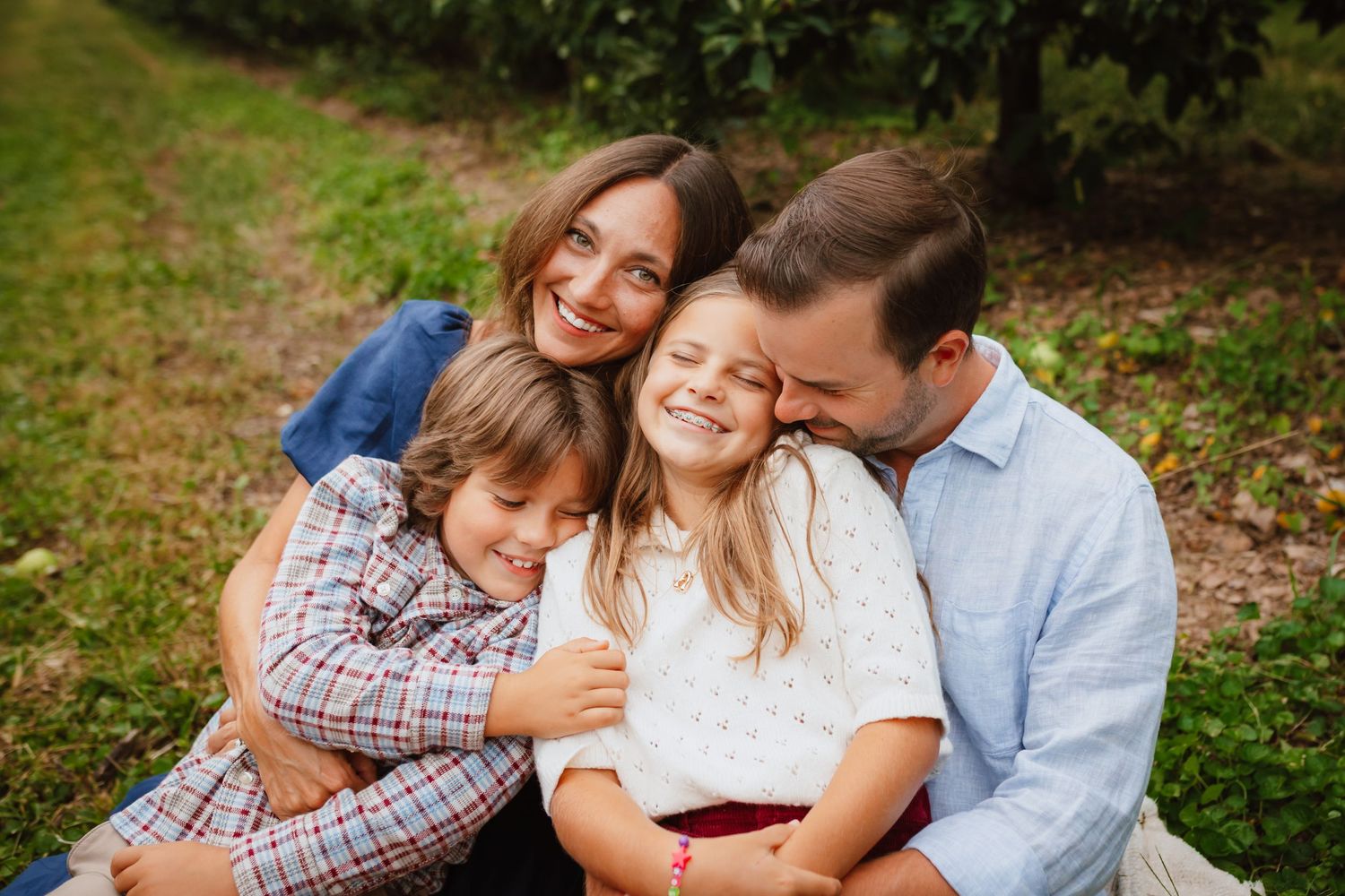 Smiling family of four hugging closely on the grass in an orchard, with parents embracing two laughing children in a warm outdoor portrait.