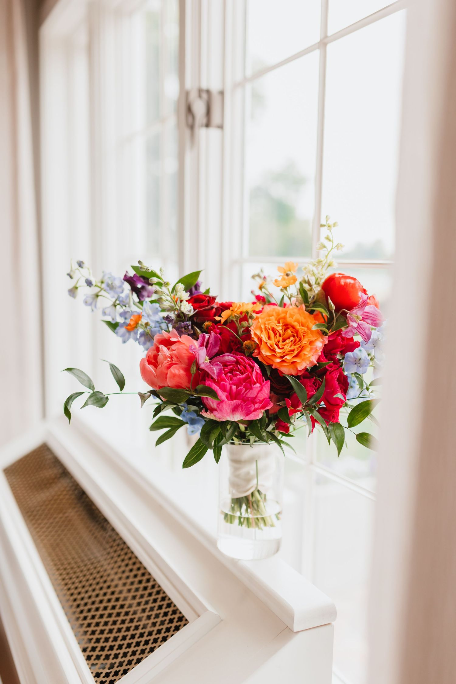 Vibrant wedding bouquet of pink peonies, orange roses, and blue flowers displayed on a bright windowsill.
