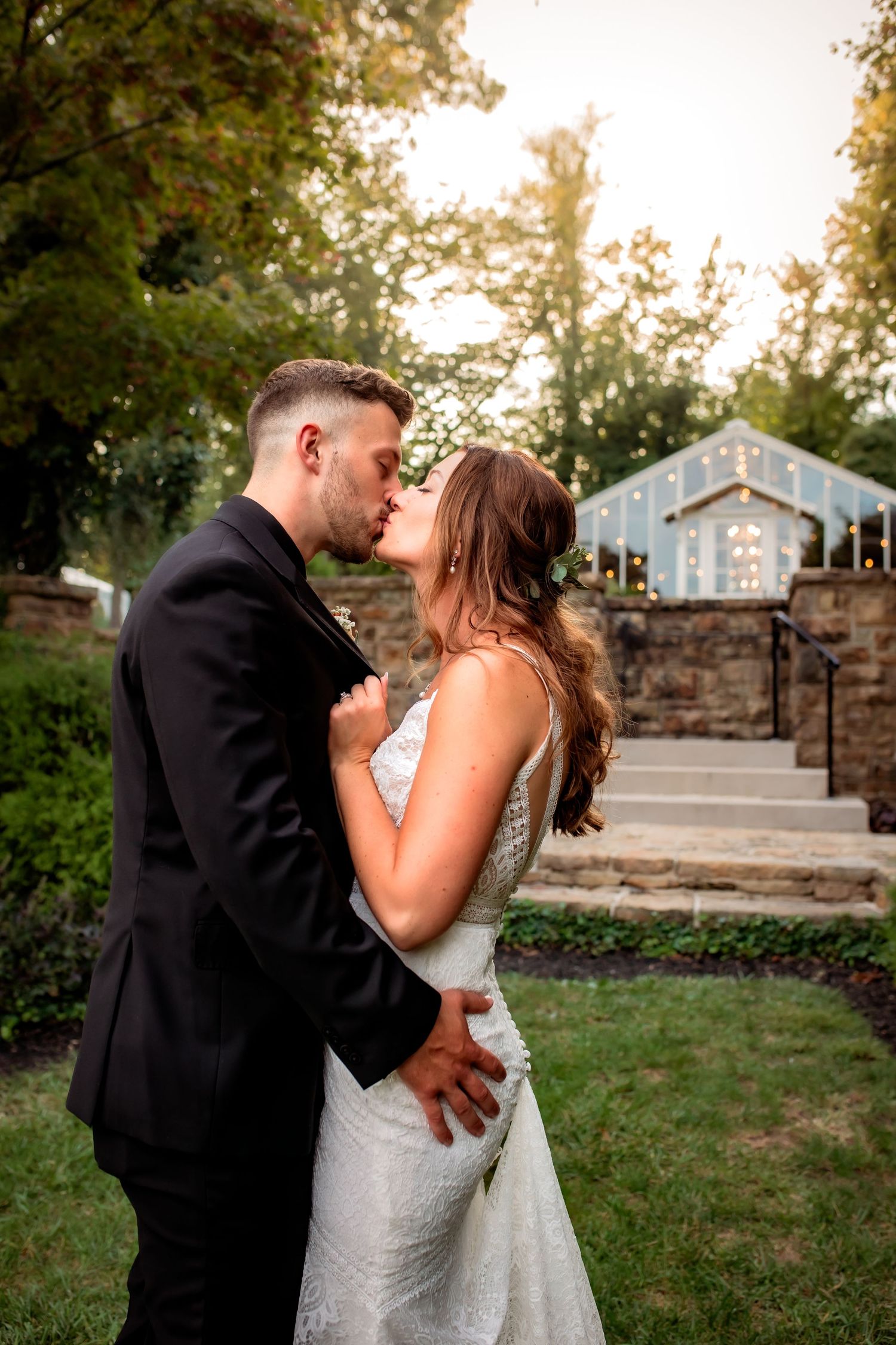 Newlyweds share a sunset kiss in a lush garden with a charming lit greenhouse in the background.