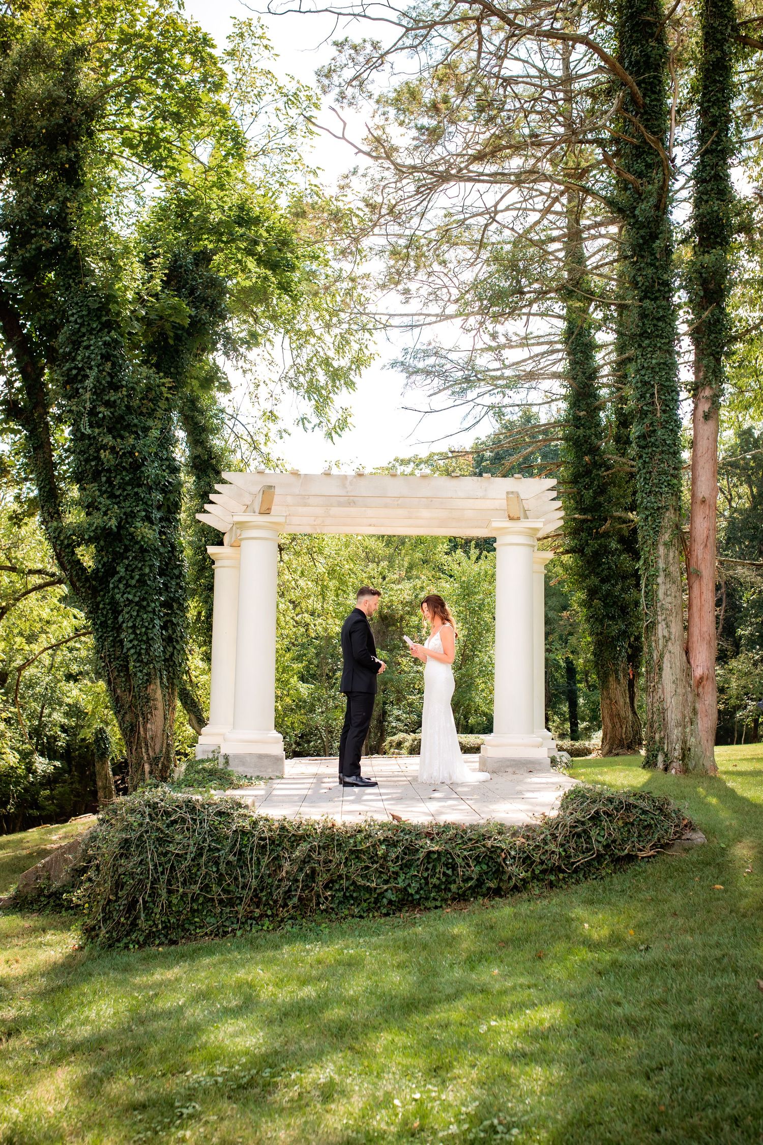 Bride and groom exchange vows under a white pergola covered in ivy surrounded by tall green trees.