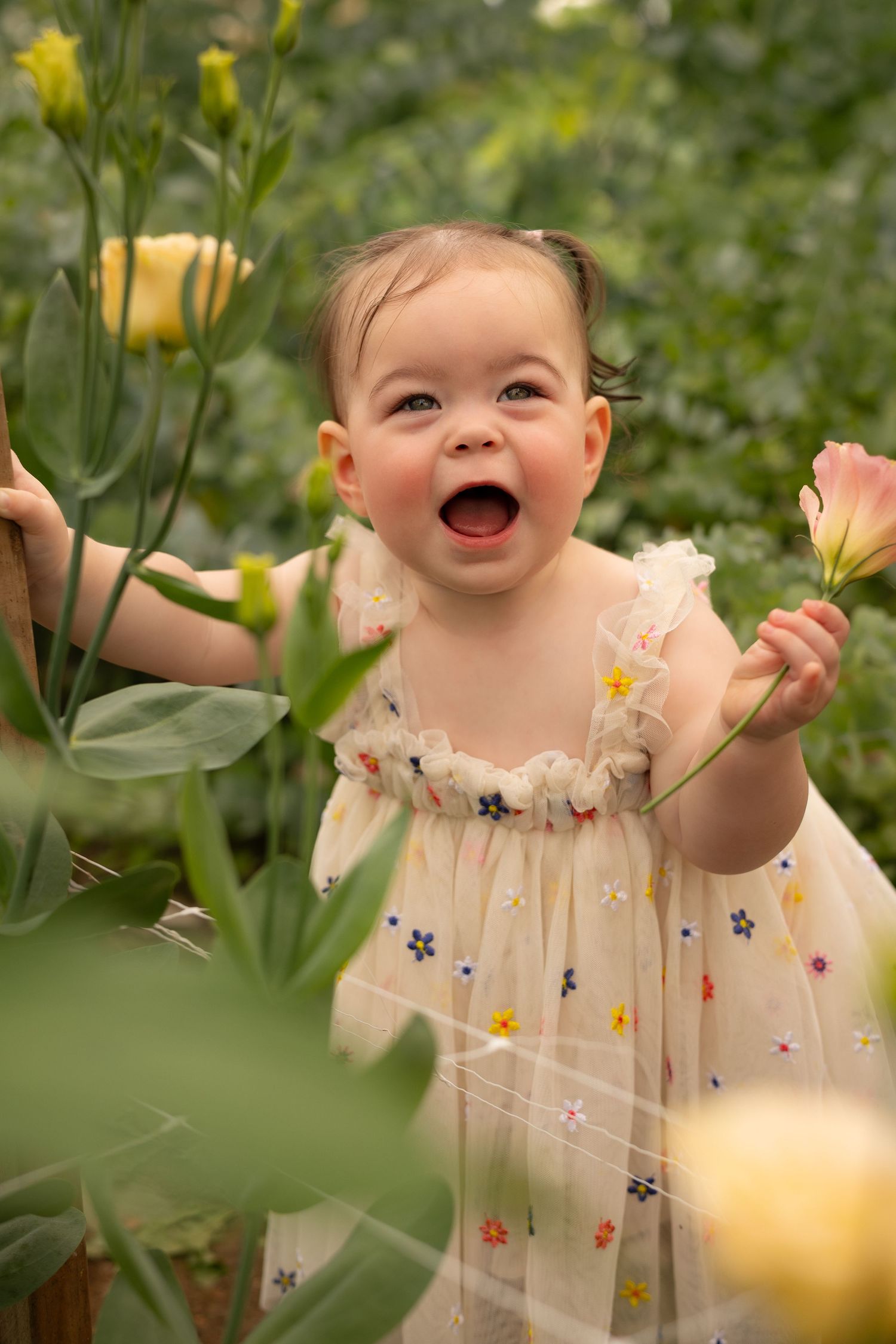Joyful baby girl in floral dress laughing among colorful garden flowers.