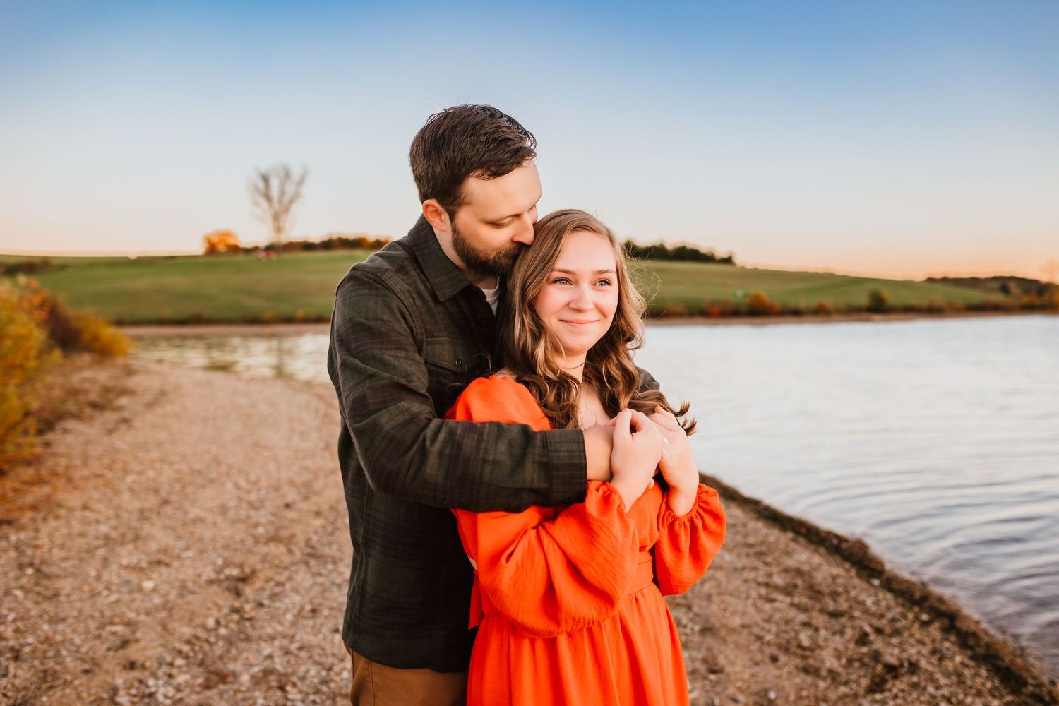 Man embracing woman in orange dress by a calm lakeside at sunset.