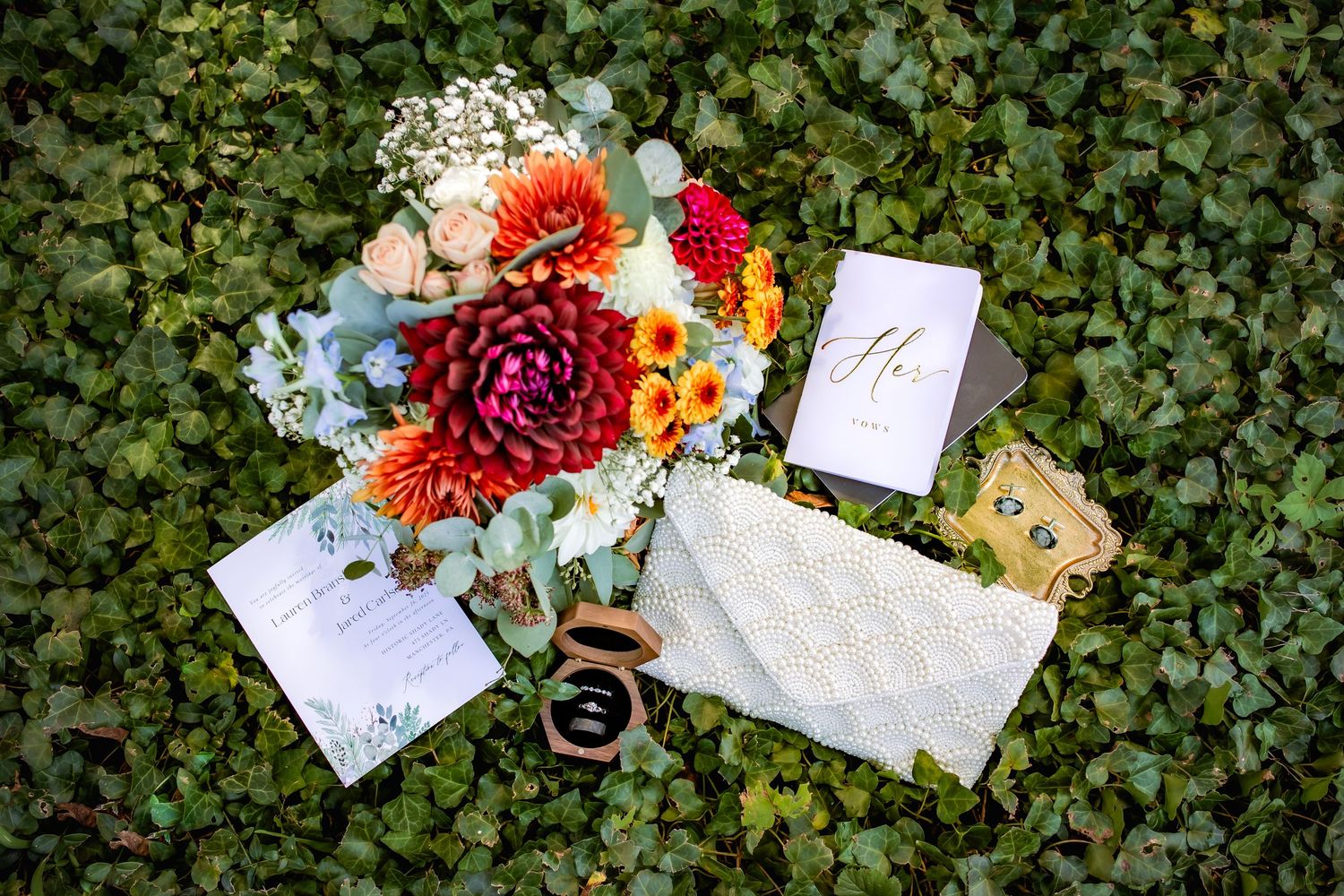 Wedding flat lay featuring colorful bouquet, invitations, rings, and accessories on green ivy.