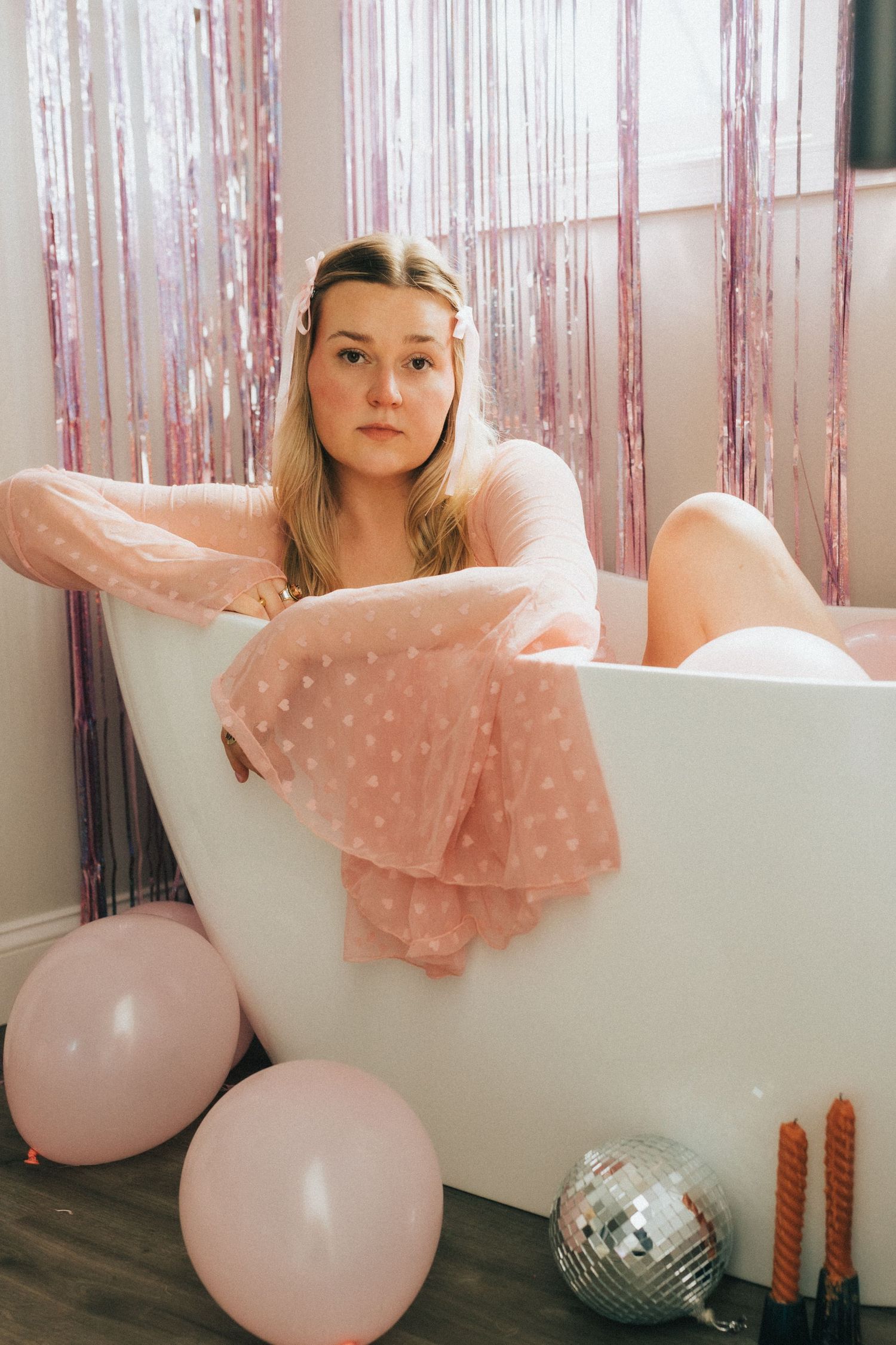 Young woman in pink polka dot dress poses in white bathtub surrounded by pink balloons and decorations.