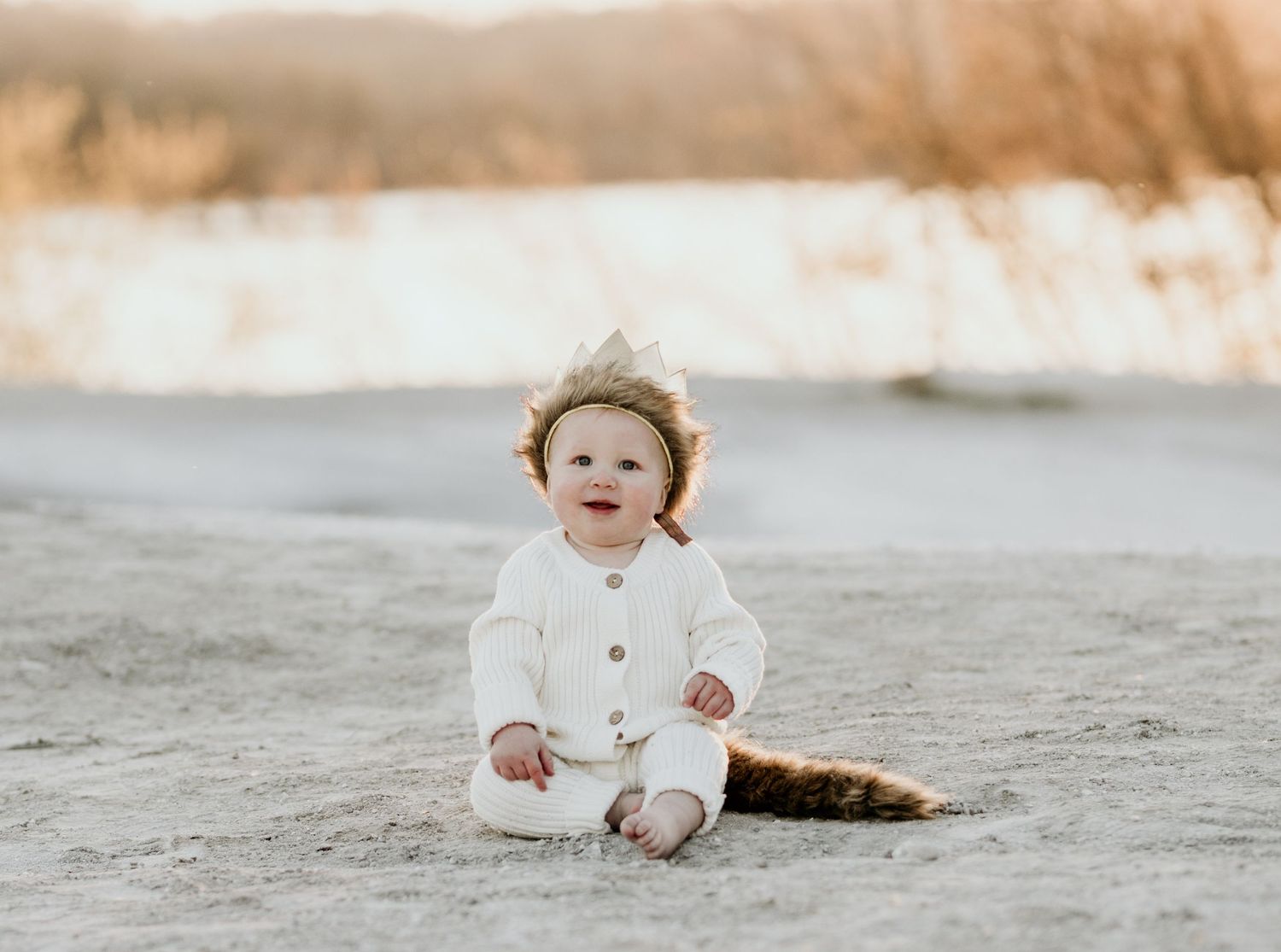Smiling baby in white knit outfit wearing crown headband sitting on sandy beach at golden hour.