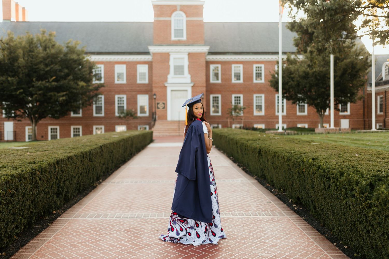 Graduate in navy cap and gown with floral skirt posing on brick path by campus building.