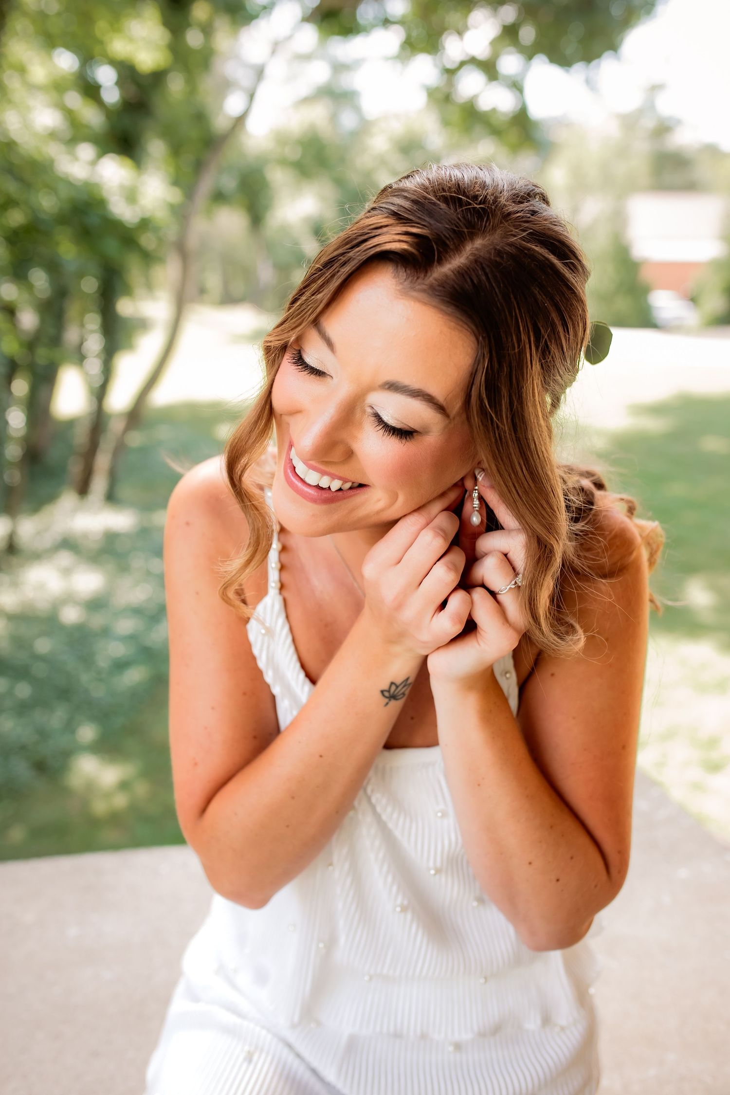 Smiling bride in white dress adjusting earring outdoors with green trees in background.