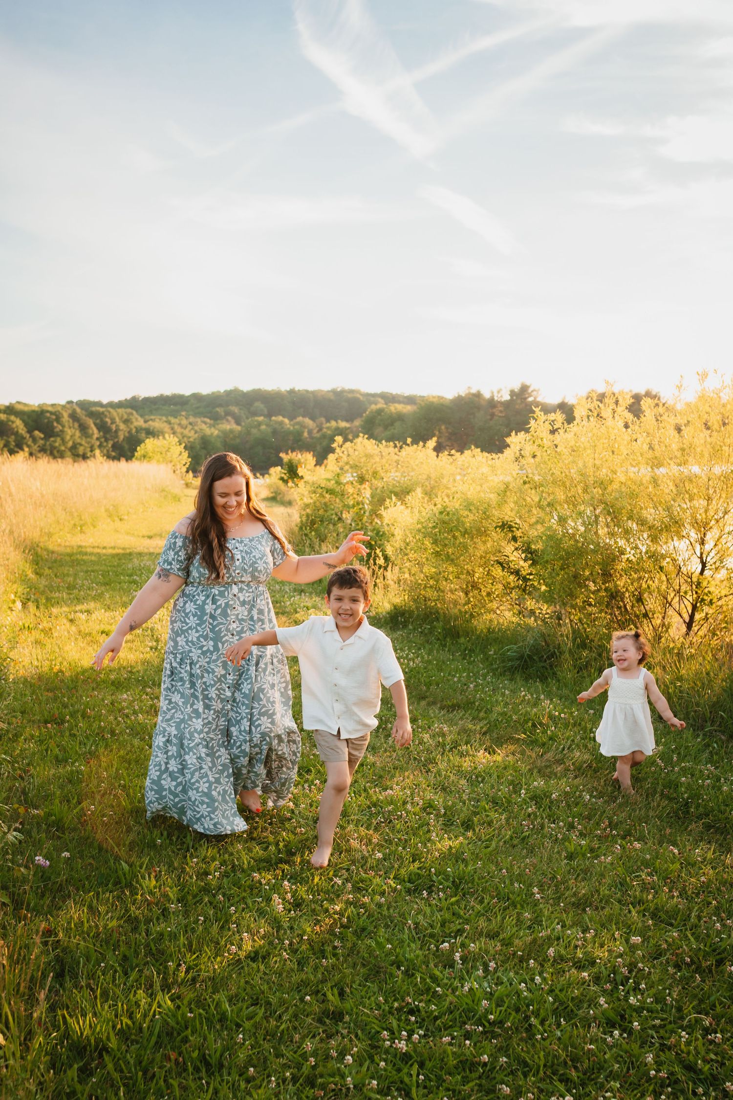 Mother walking barefoot through golden meadow with two young children at sunset in summer.
