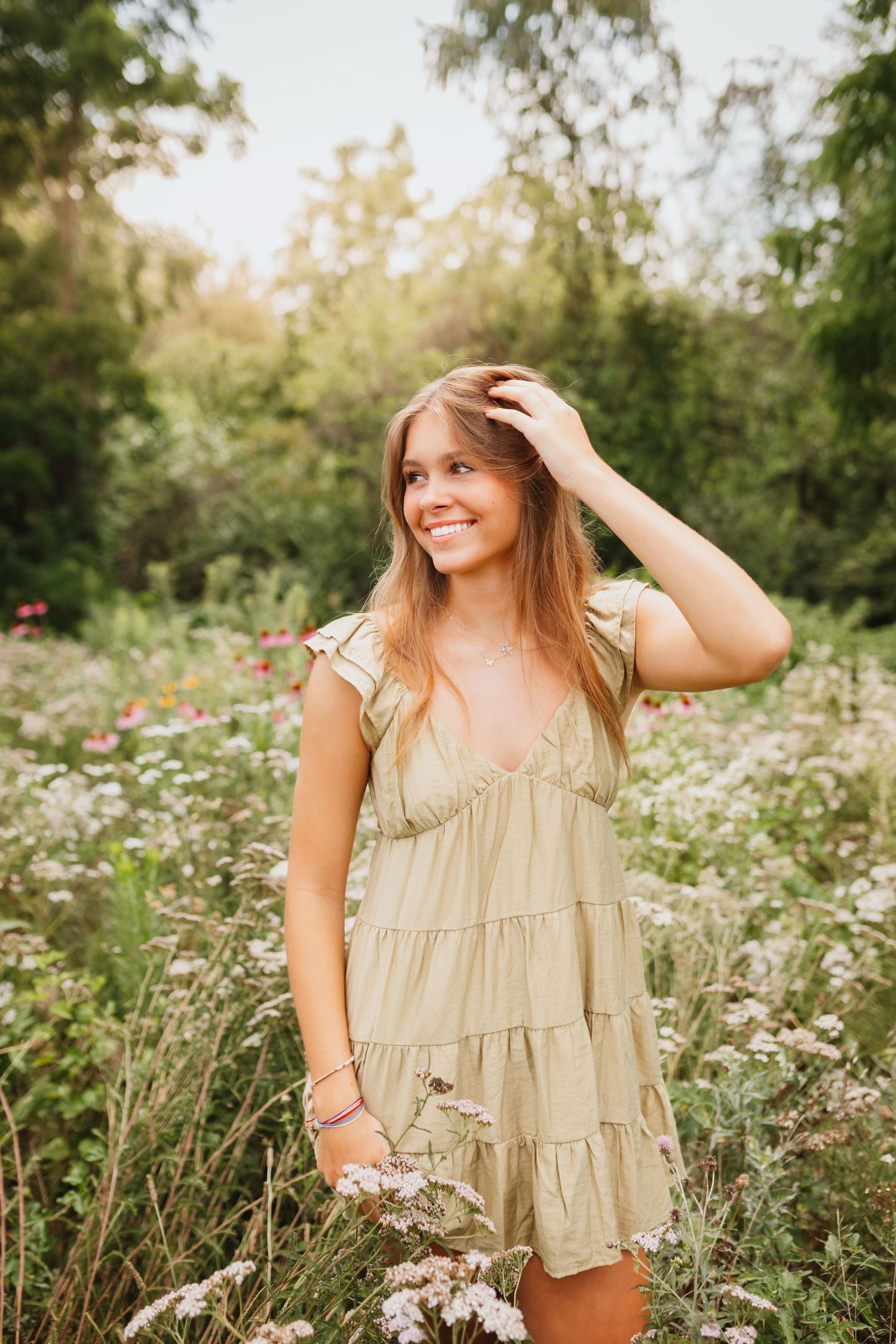 Smiling teenage girl in a sage green dress standing among wildflowers in a sunny garden.