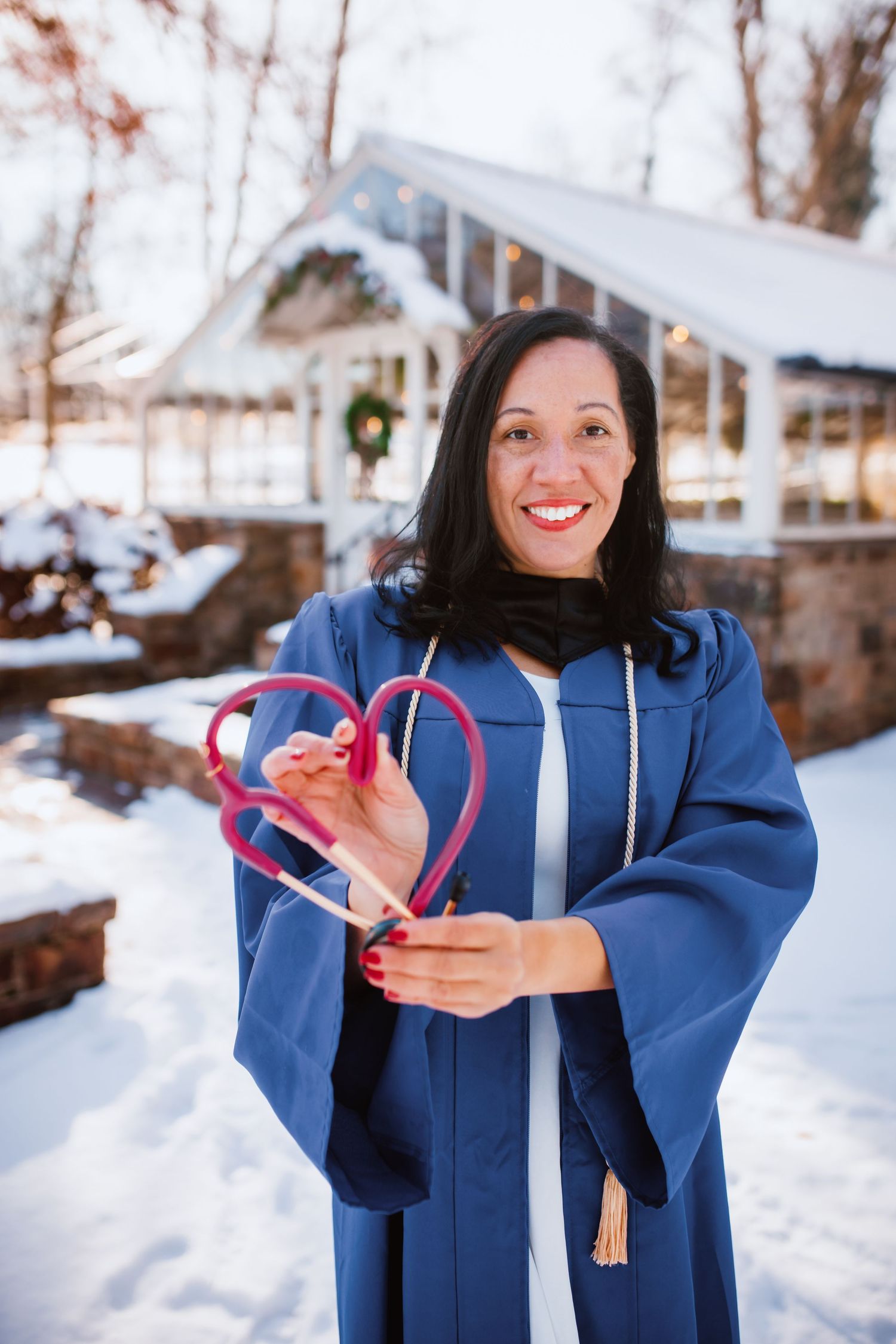 Graduate in blue academic gown holds pink stethoscope heart in snowy outdoor setting.