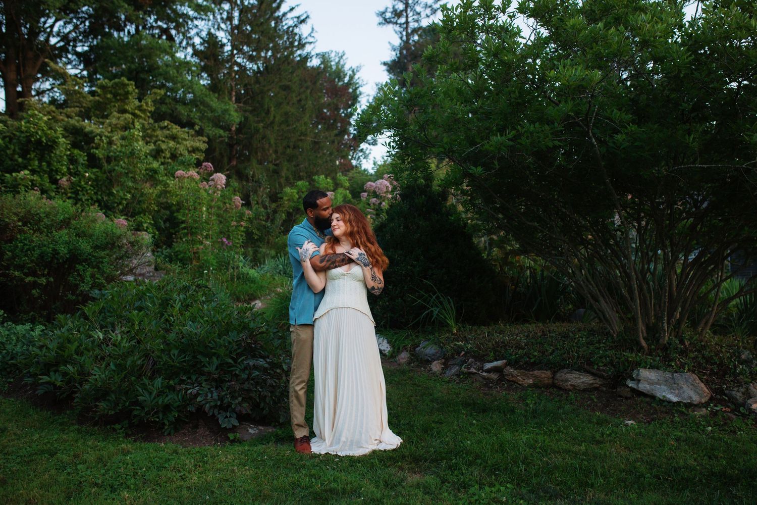 Romantic couple embracing in a lush green garden surrounded by blooming flowers at dusk.