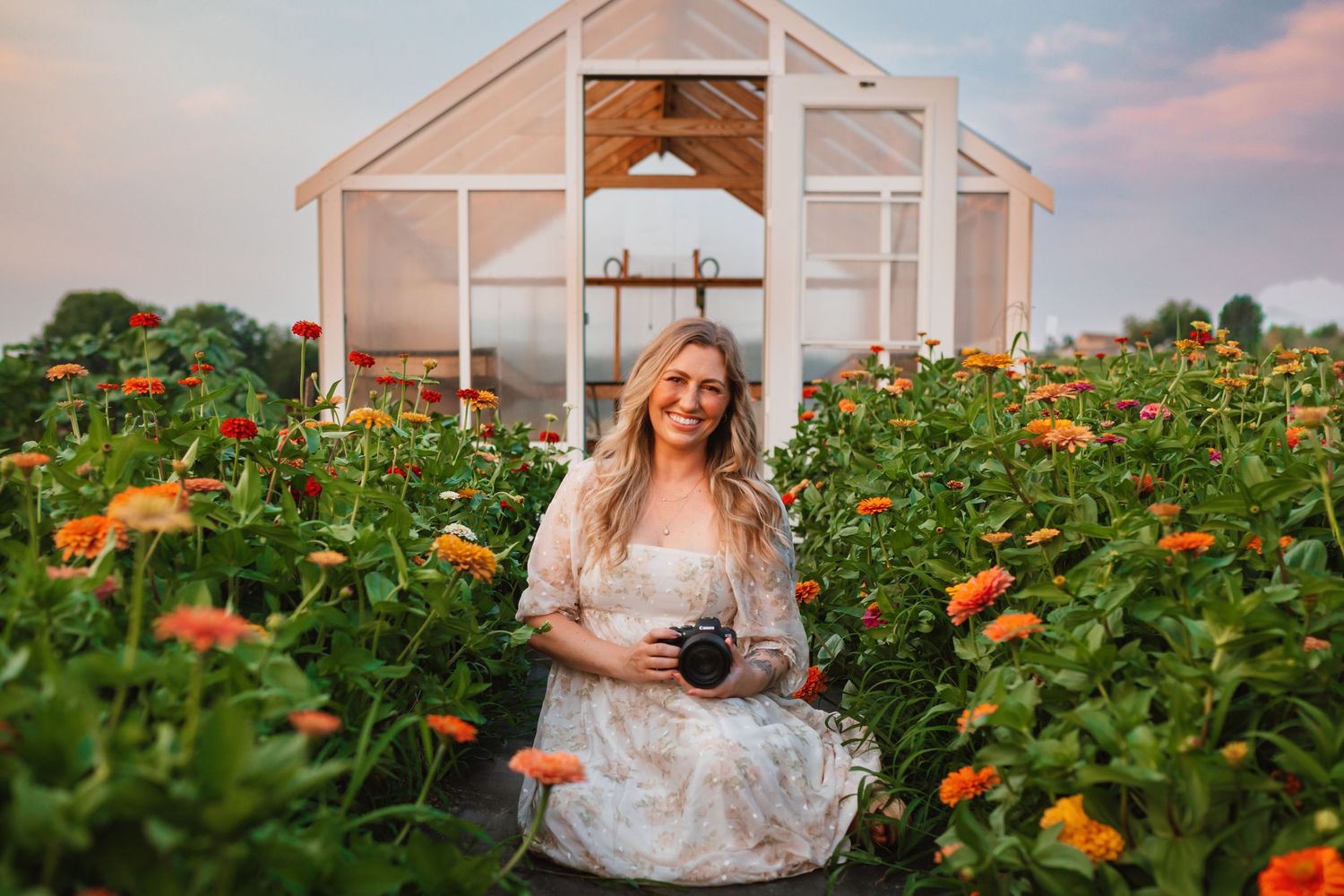 Smiling blonde photographer sits among vibrant orange zinnias in front of a white greenhouse at sunset.