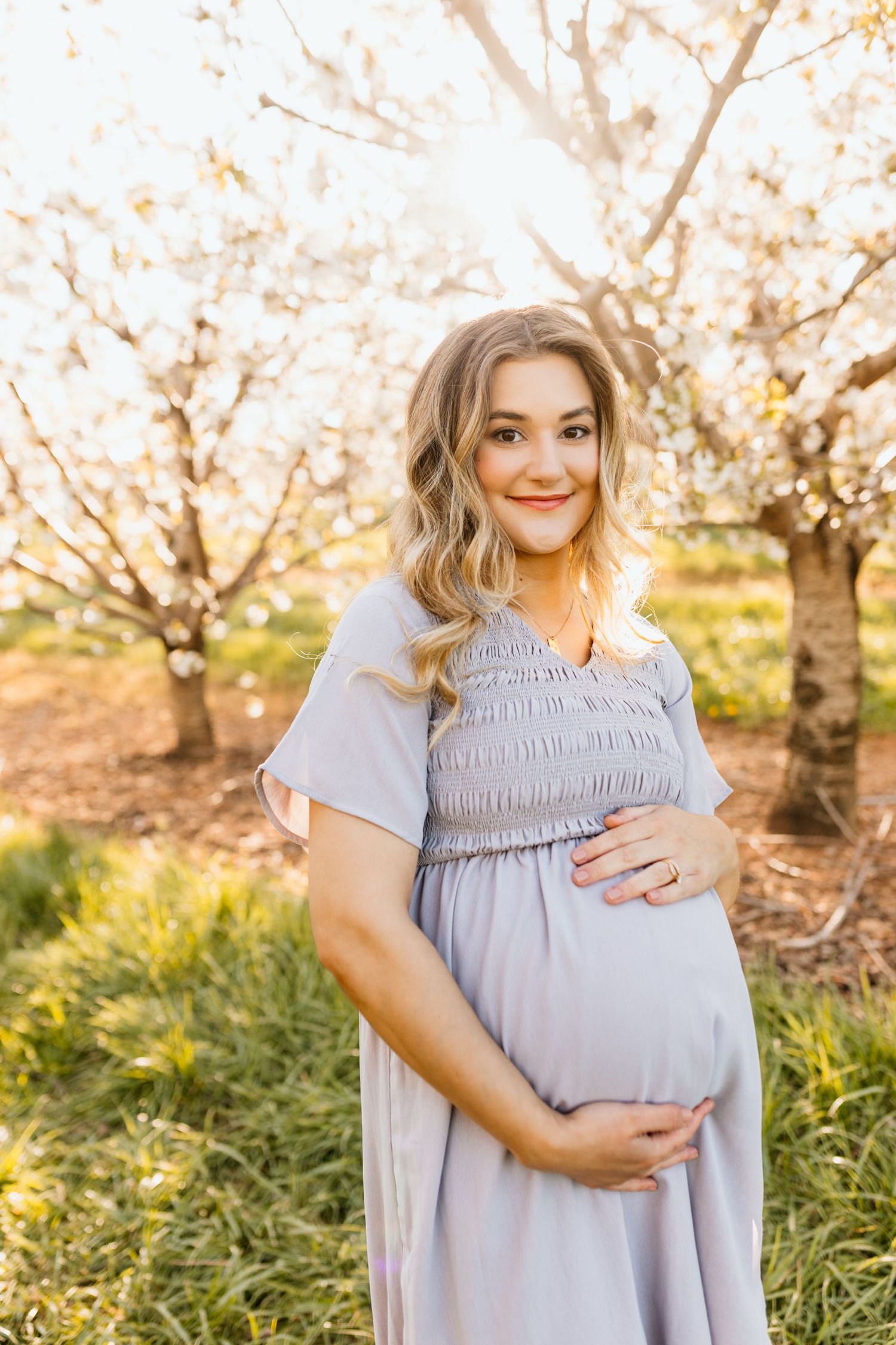 A smiling pregnant woman in a lavender dress cradling her baby bump among blooming spring trees.