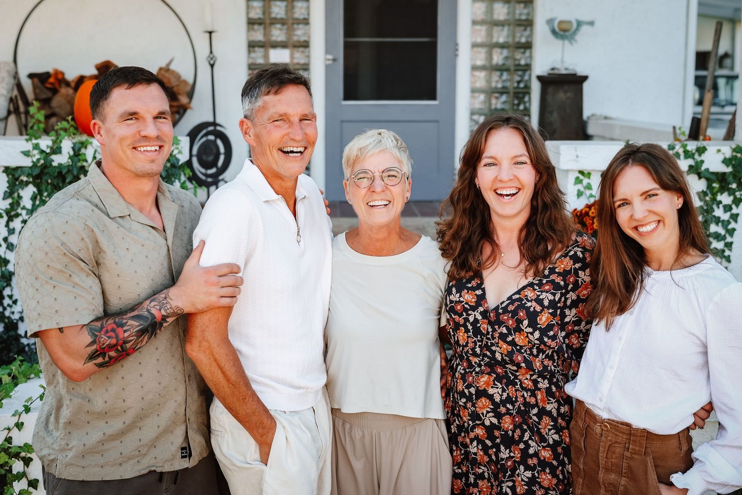 Five smiling family members pose together outdoors in front of a white home.