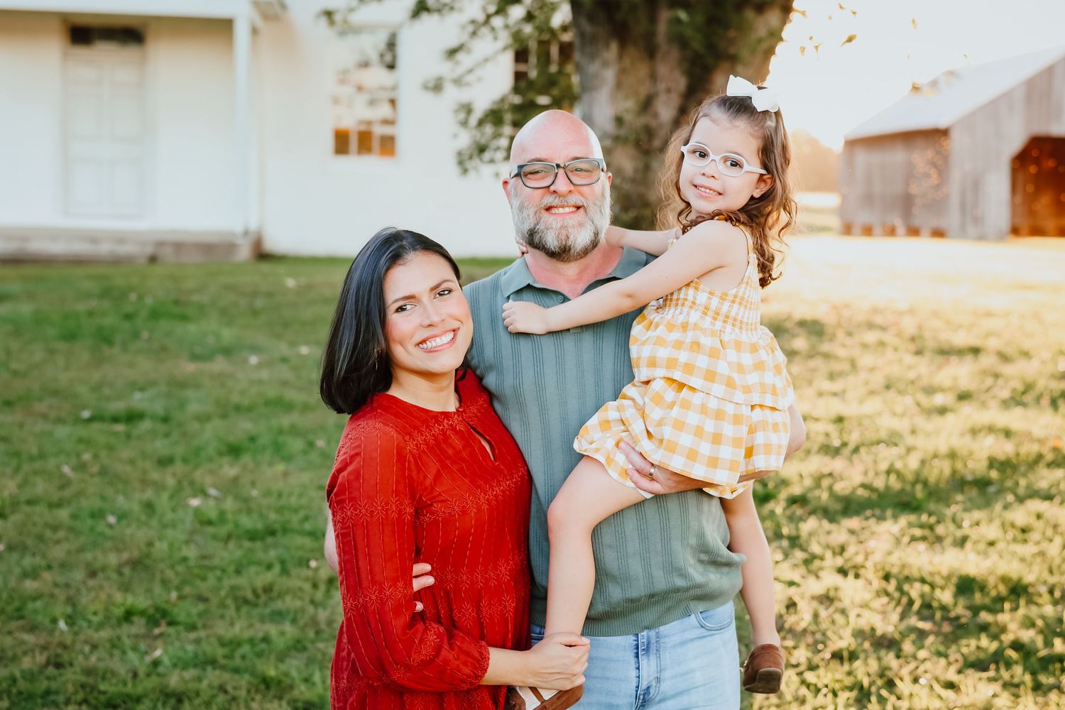 Smiling family of three with young daughter posing outdoors near a white building.
