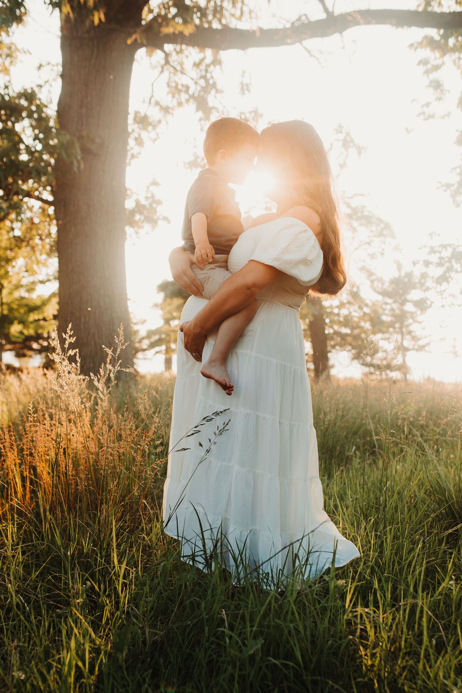 Mother holding child in golden sunset light wearing white dress in grassy field near tree.