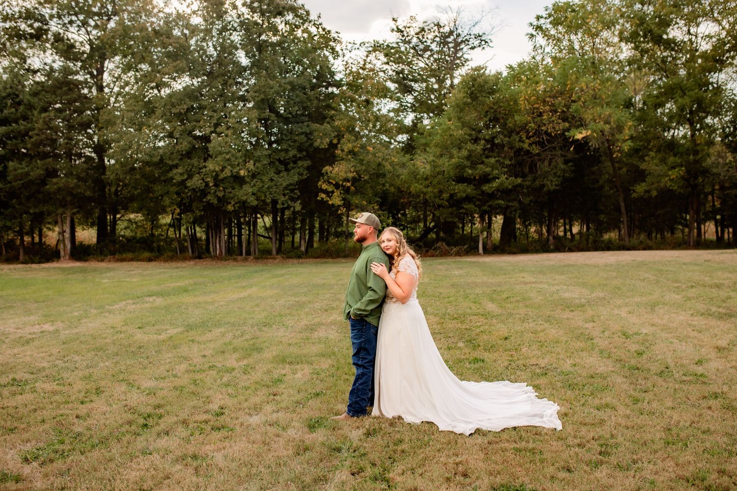 Bride and groom stand together in an open green field surrounded by trees during their outdoor wedding portrait.