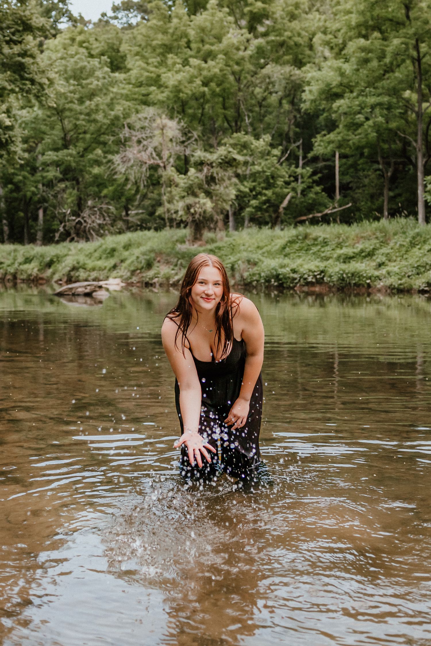 Smiling young woman in black dress splashing in a shallow creek surrounded by green trees.