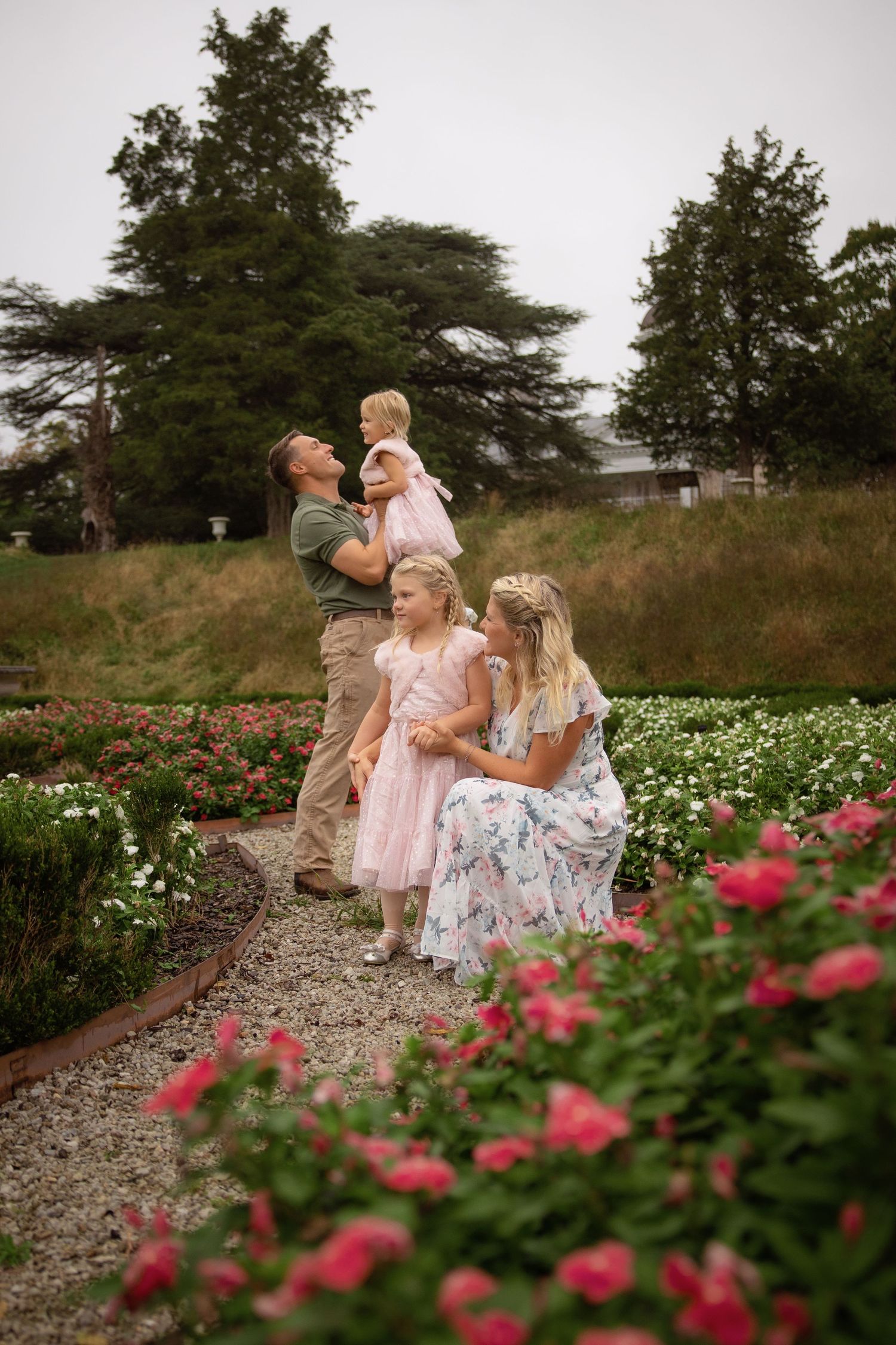 Family of four in a rose garden, a father holding a toddler while a mother in a floral dress sits beside a girl in a pink tulle dress.