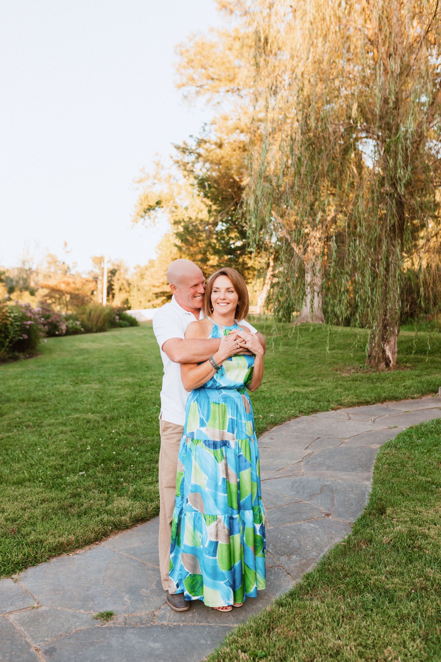 Man embracing woman in colorful maxi dress on garden path under willow tree.