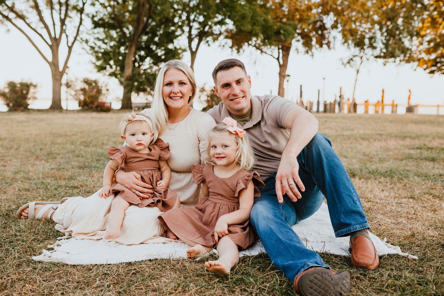 A smiling family of four sits on a blanket in a sunny park during fall.