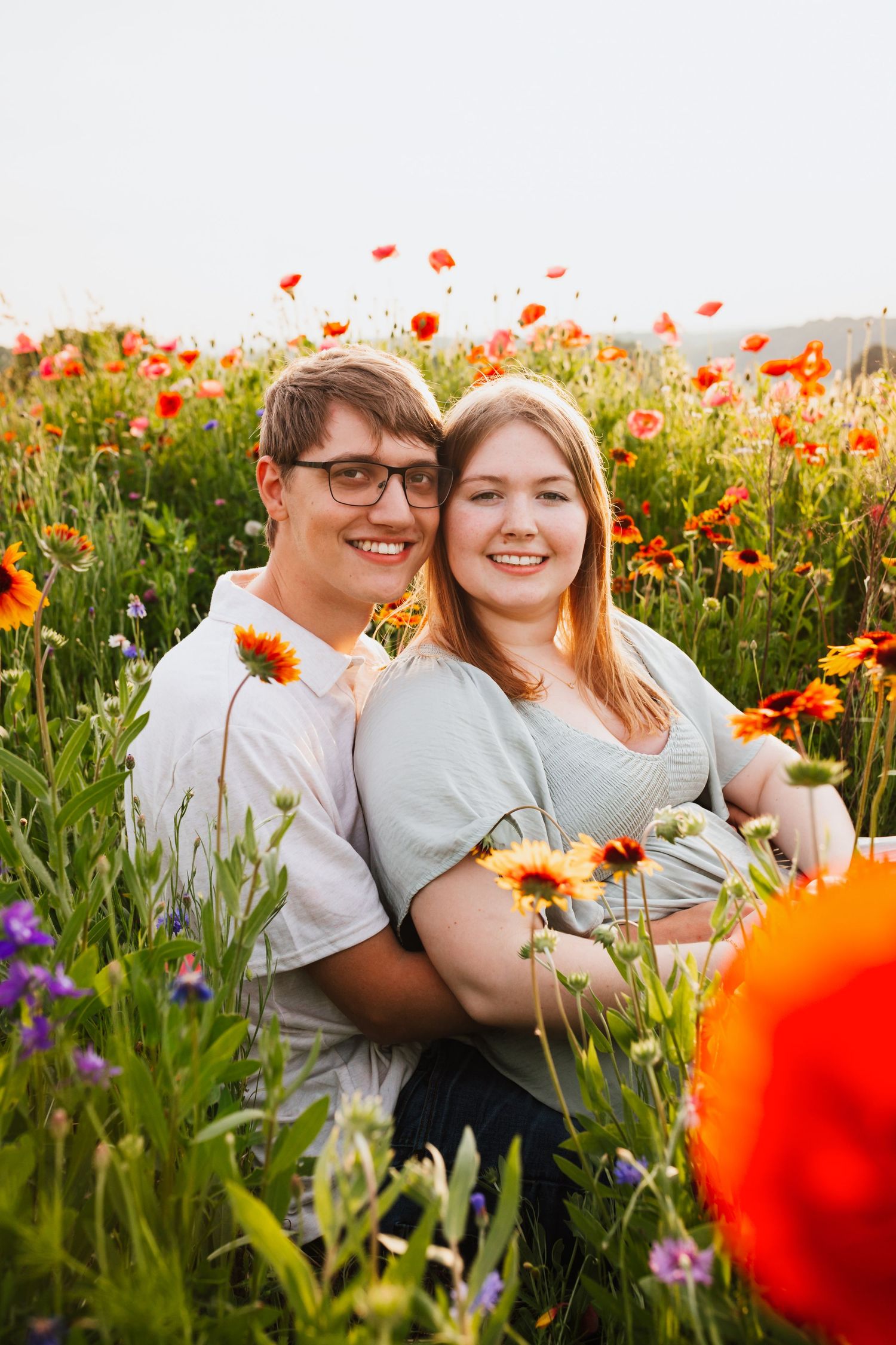 Smiling couple sitting together in a vibrant wildflower field filled with red poppies and colorful blooms.